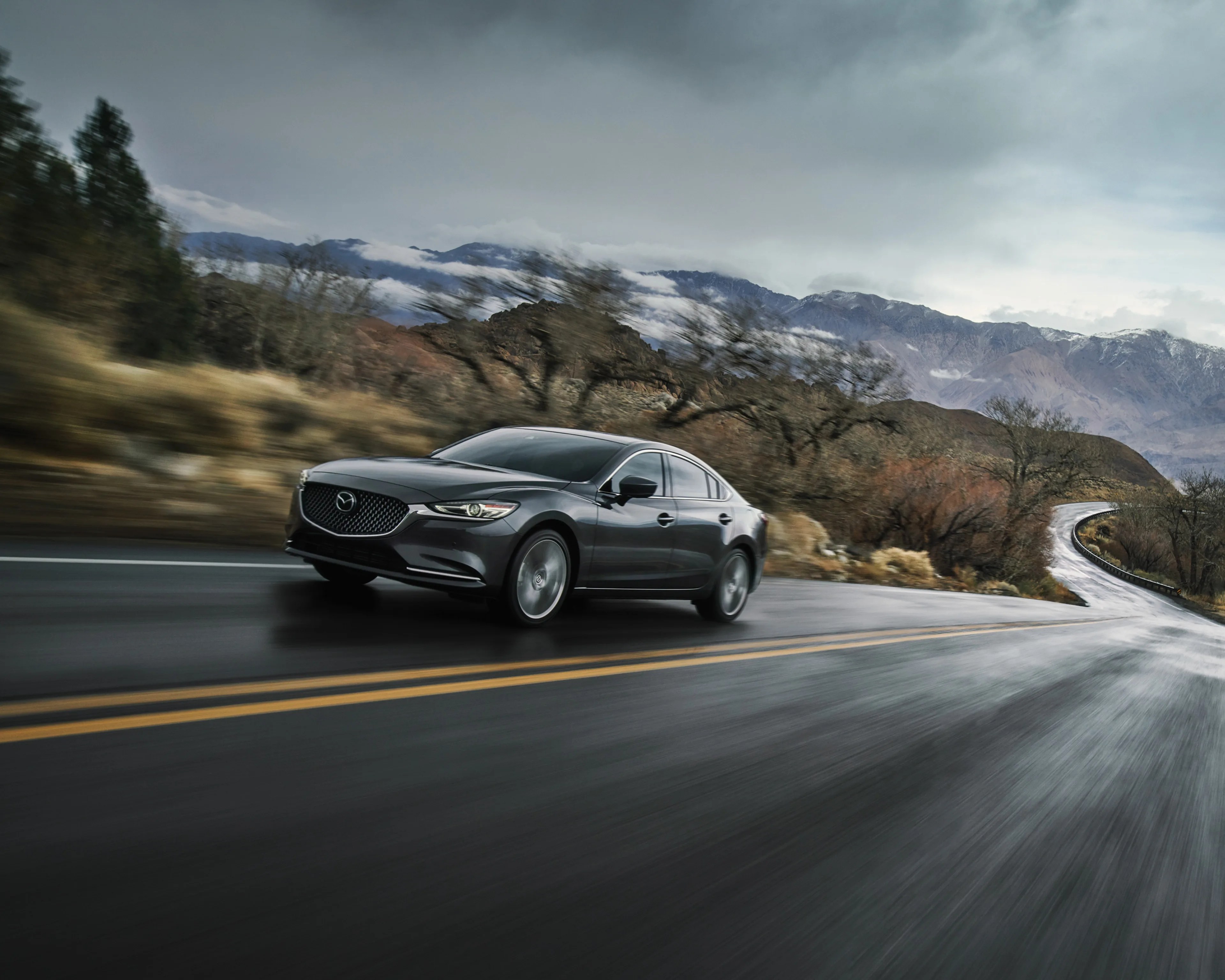 Dark gray Mazda sedan driving on a wet winding road with mountains and cloudy sky in the background.