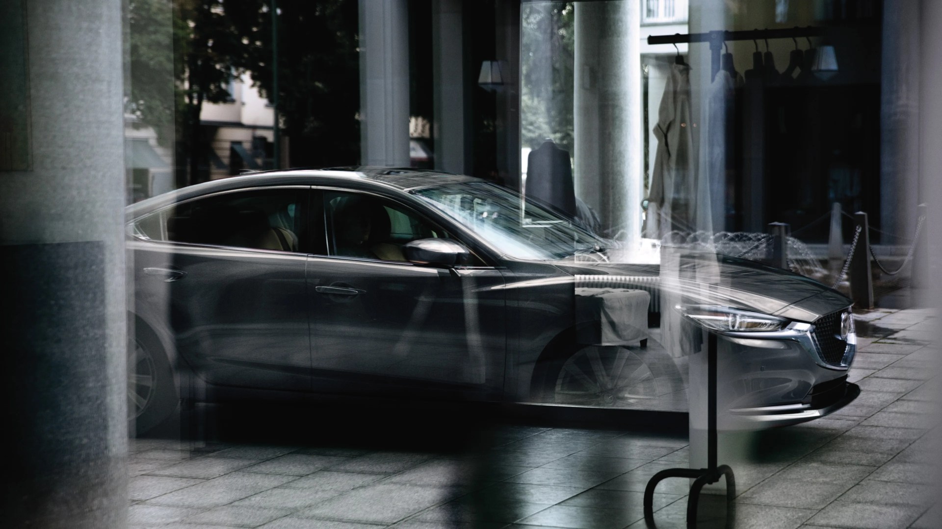 Dark gray sedan parked indoors with reflections of clothing racks and columns on the glass.