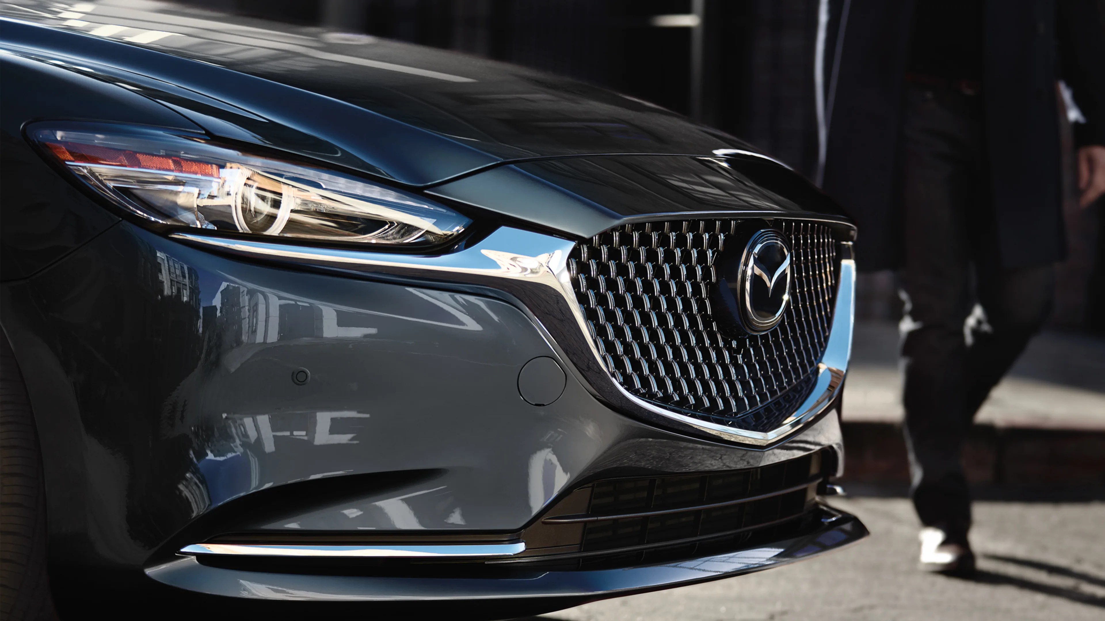 Front close-up of a dark gray Mazda car showing the headlight, grille with Mazda logo, and part of a person walking nearby.
