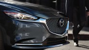 Front close-up of a dark gray Mazda car showing the headlight, grille with Mazda logo, and part of a person walking nearby.