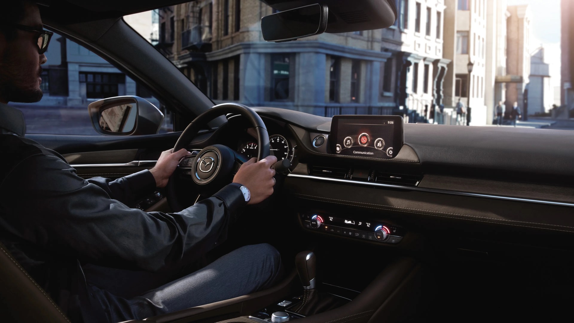Driver holding a black leather-wrapped Mazda steering wheel inside a car with digital display and climate controls.