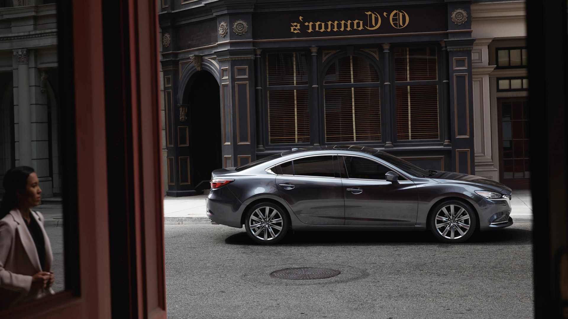 Gray sedan parked on a city street in front of a black and gold ornate building with a woman in a beige blazer reflected in a window.