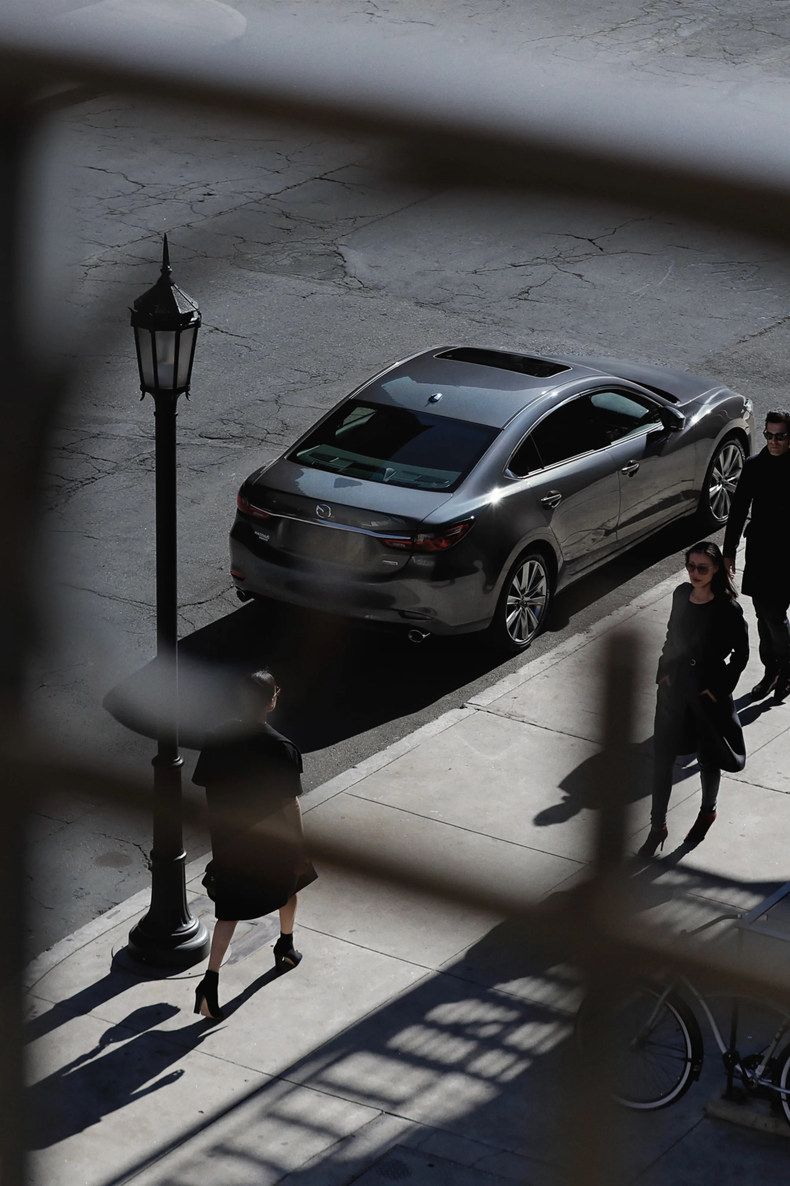 Gray Mazda sedan parked on a street next to a sidewalk with three people walking nearby.