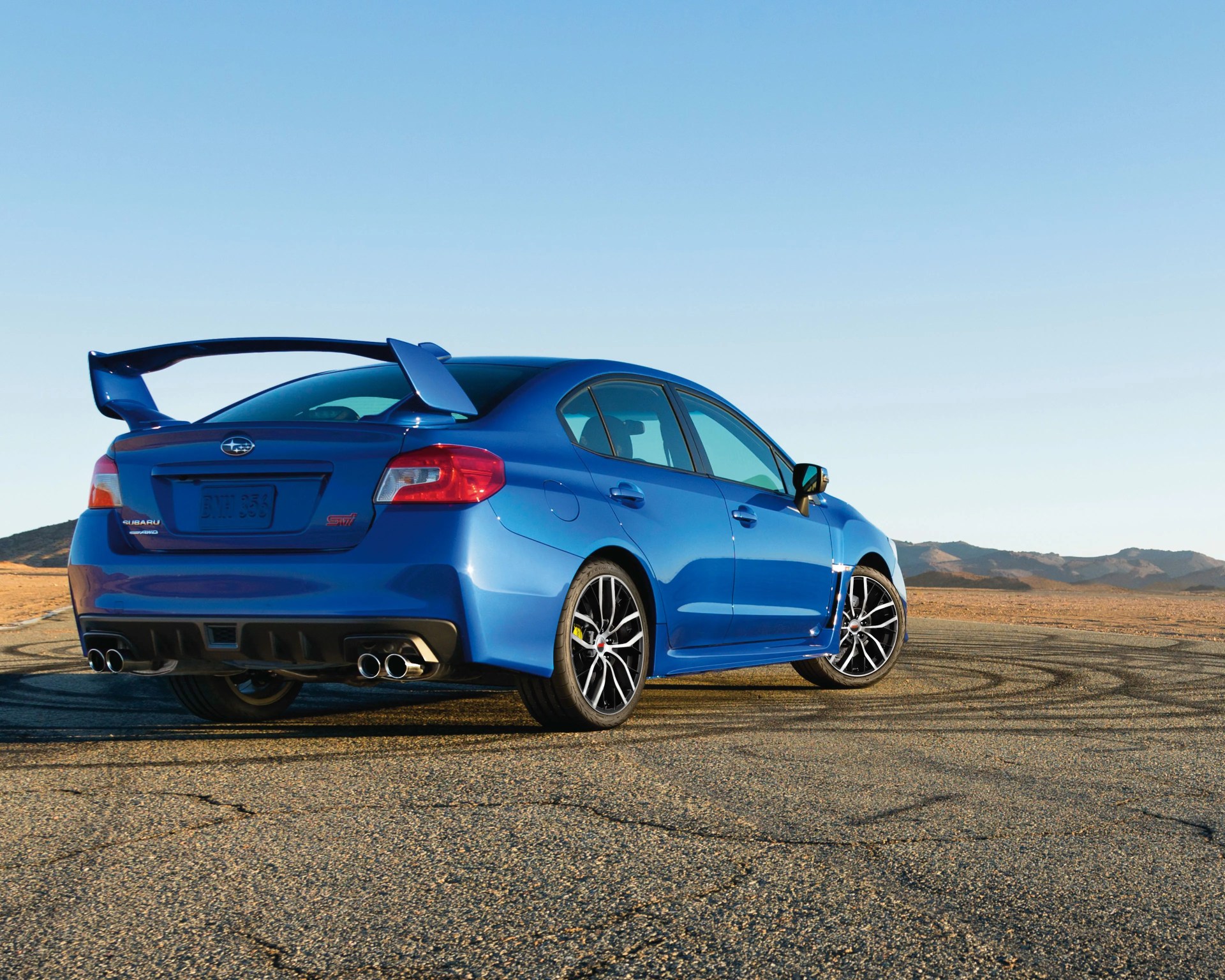 Blue Subaru WRX STI sedan with large rear spoiler and quad exhausts on a paved desert road.