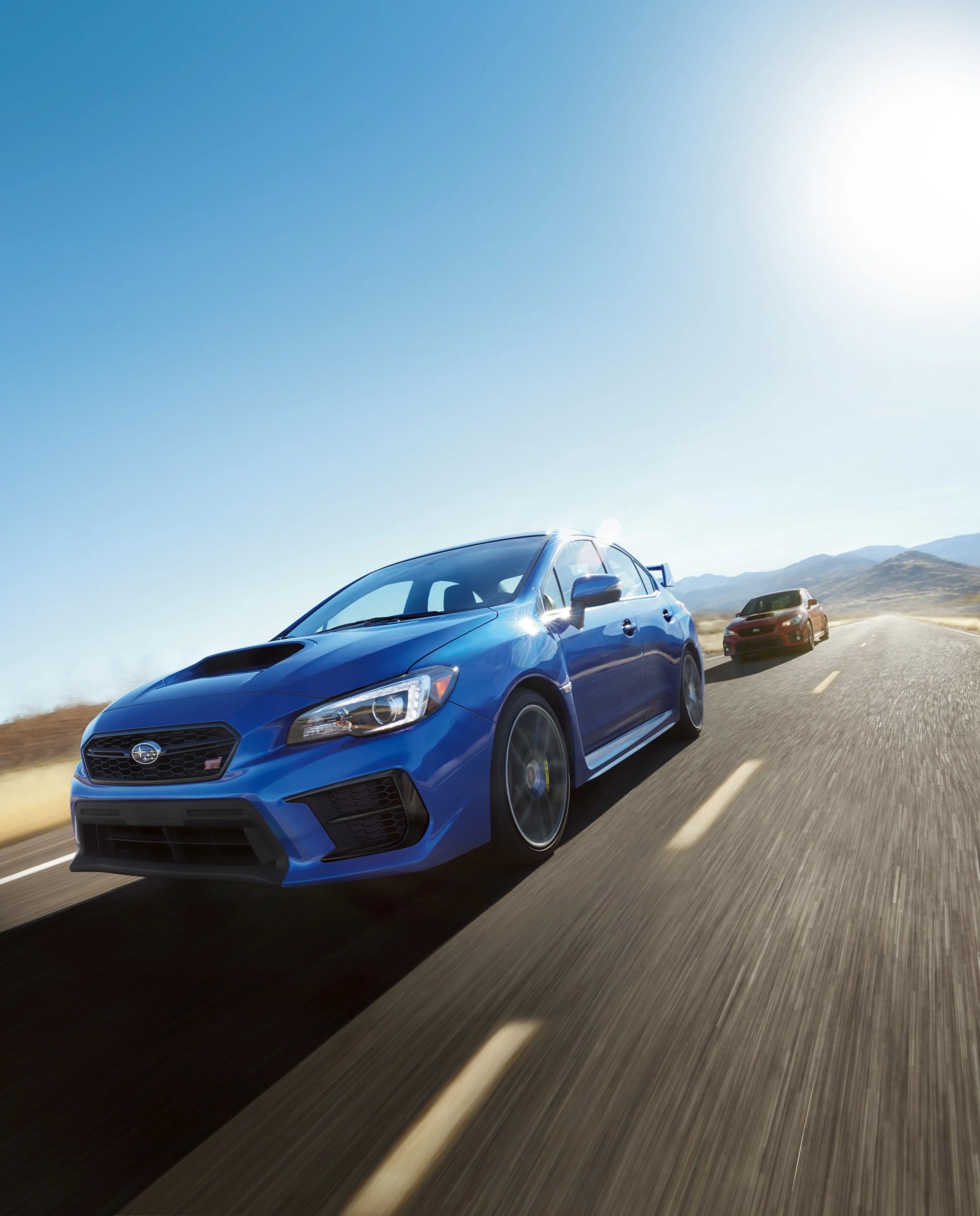 Blue Subaru WRX STI driving on a sunny open road with a red Subaru WRX STI behind.