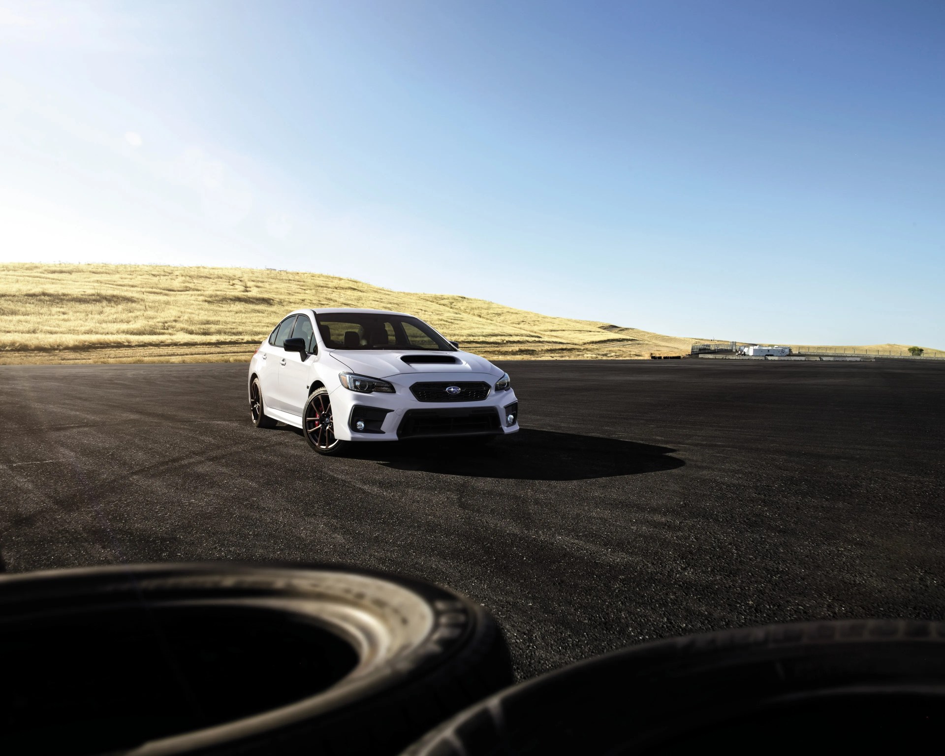 White Subaru WRX sedan parked on a large asphalt surface with dry grassy hills in the background.