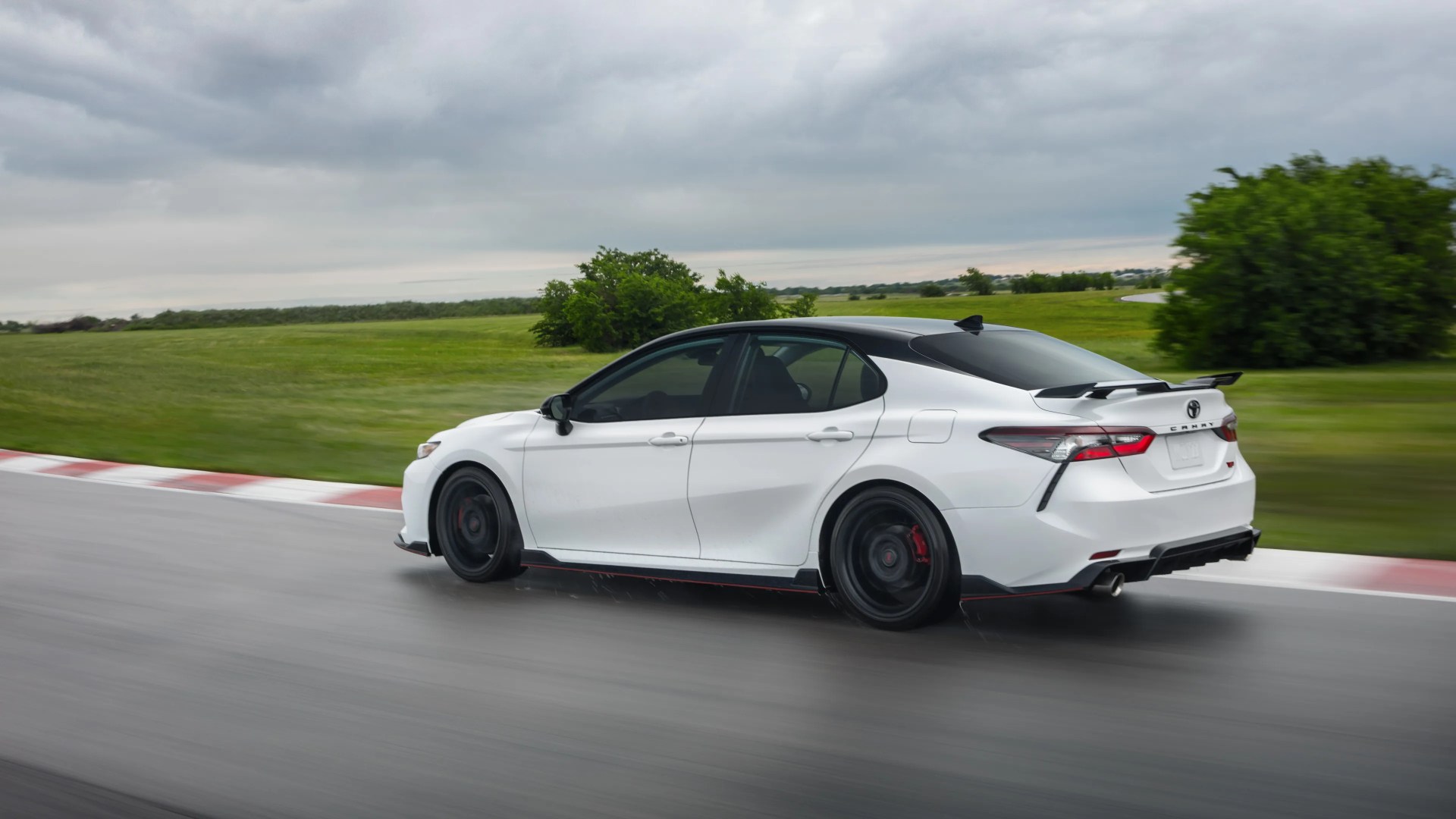White Toyota Camry sedan with black roof and spoiler driving on a wet race track with green grass and cloudy sky.
