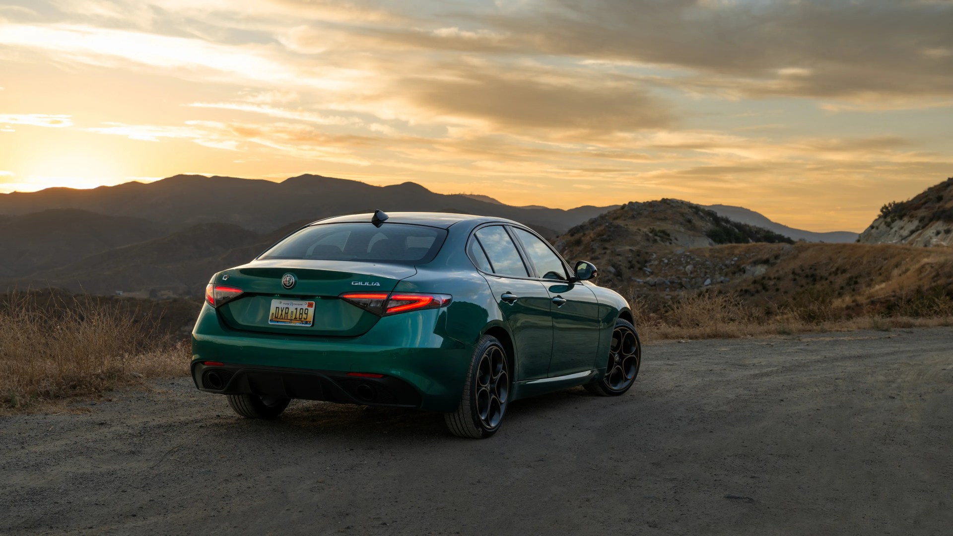 Dark green Alfa Romeo Giulia sedan parked on a dirt road with mountains and sunset in the background.