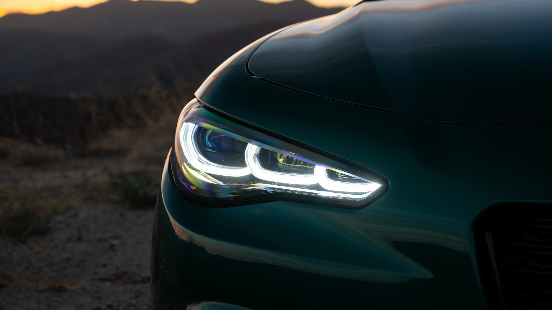 Close-up of a dark green car's LED headlight illuminated at dusk with mountains in the background.