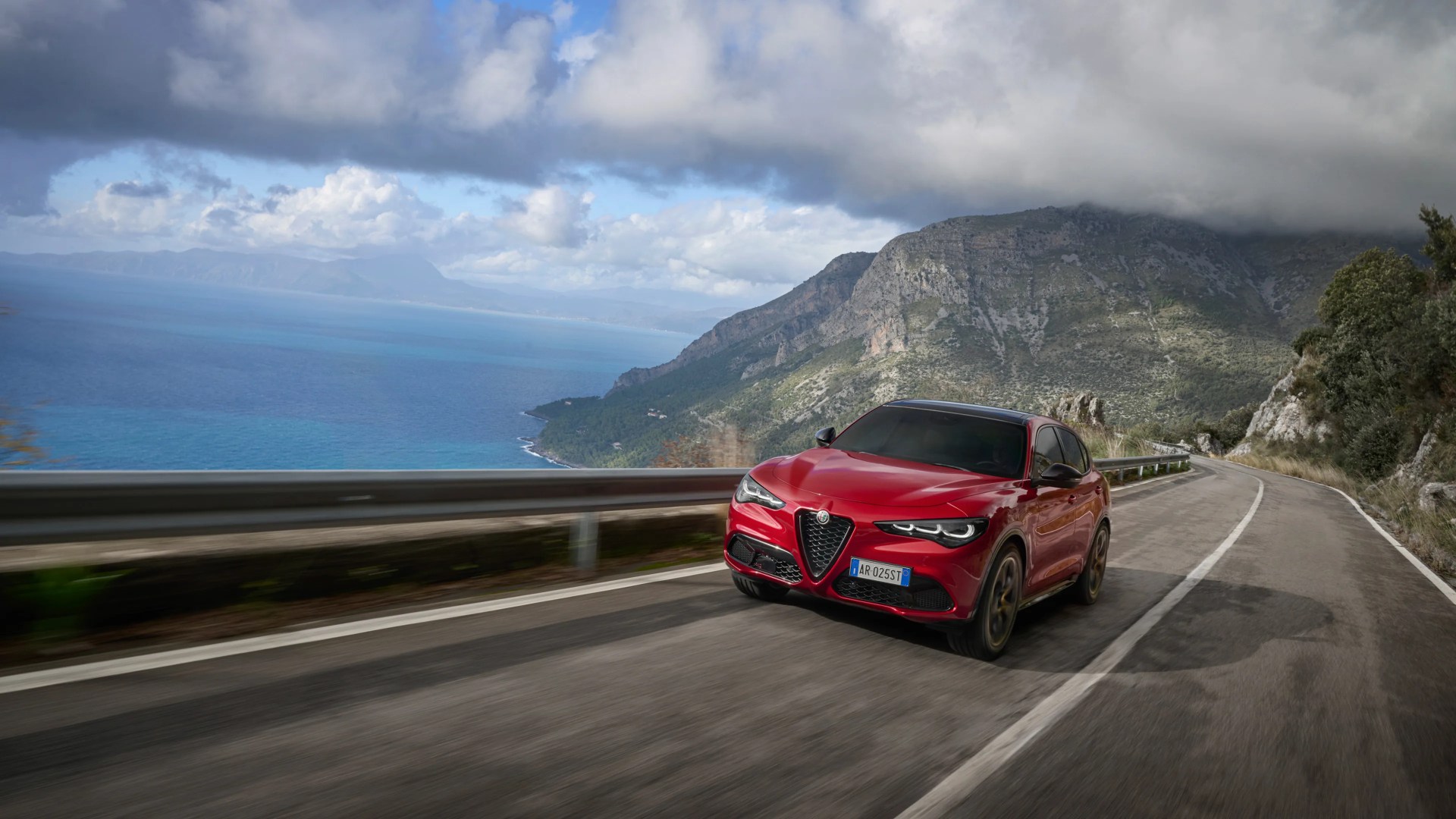 Red Alfa Romeo SUV driving on a coastal mountain road under a cloudy sky.