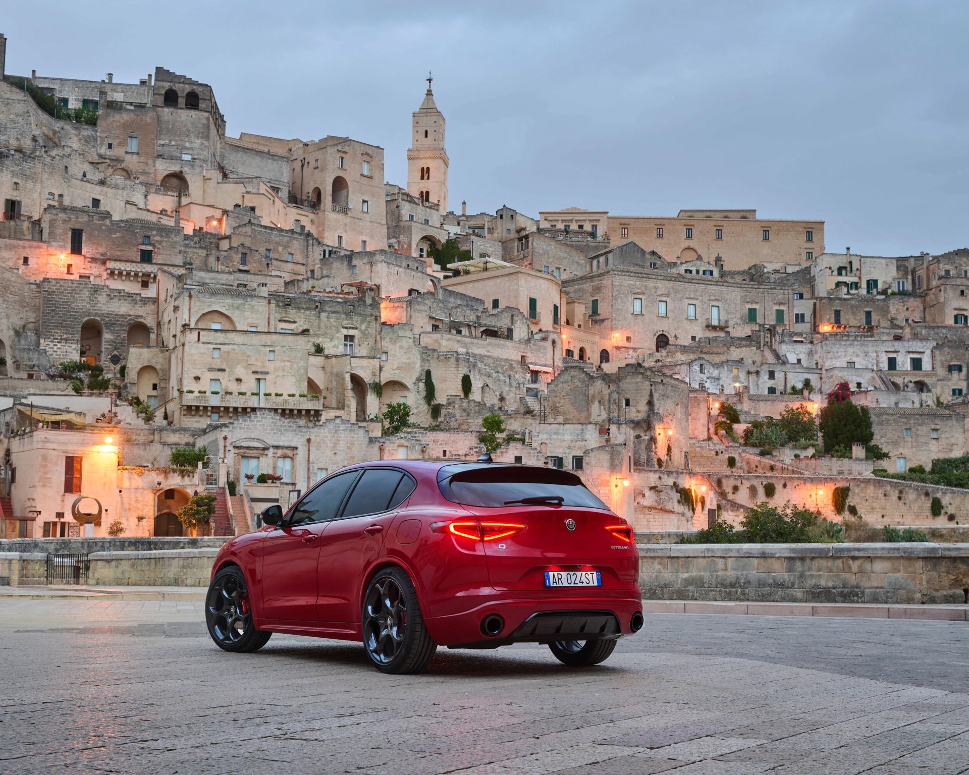 Red Alfa Romeo Stelvio SUV with rear lights on parked in front of historic stone buildings at dusk.
