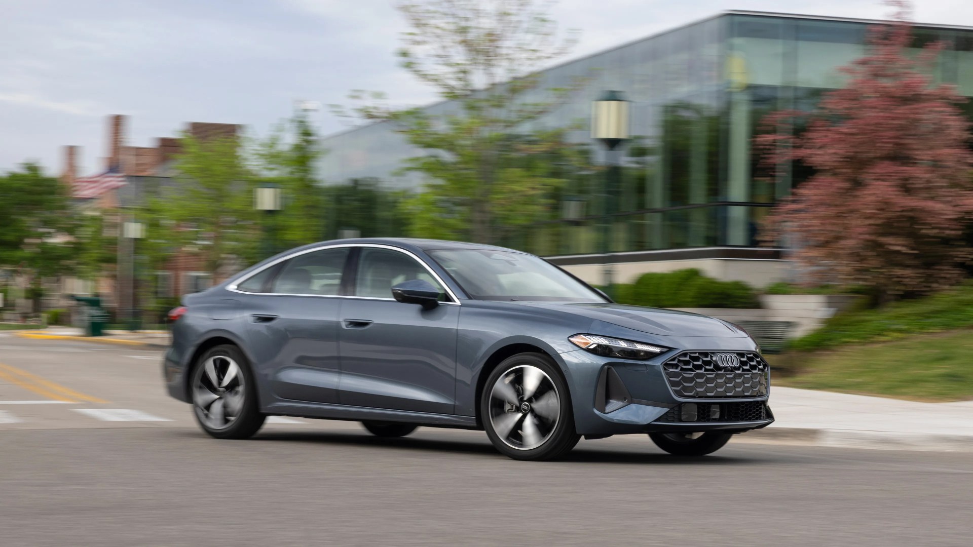 Gray Audi sedan driving on a street with modern glass building and trees in the background.