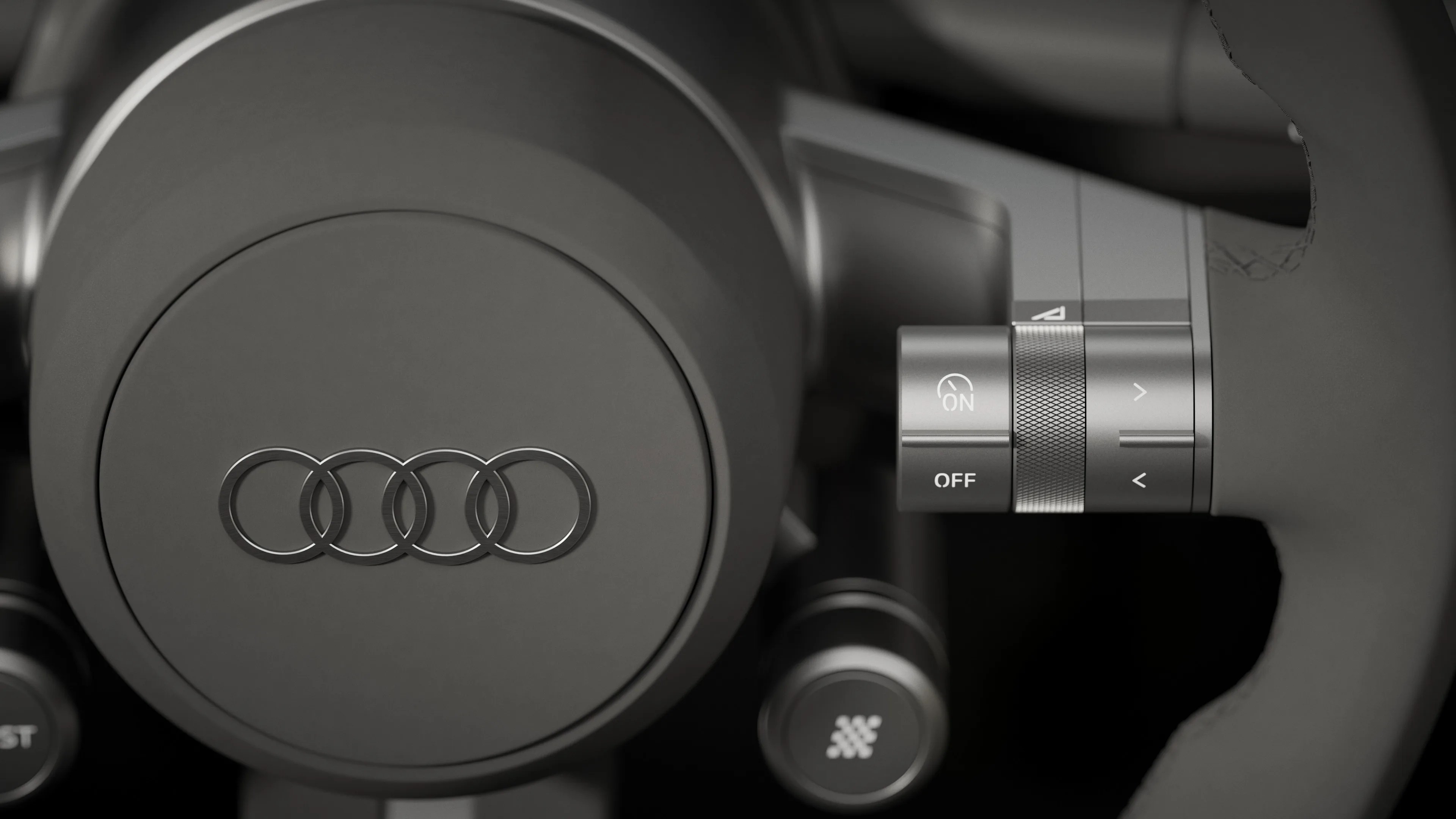 Close-up of a matte black Audi steering wheel center with four-ring logo and adjacent control knob with ON/OFF and arrow buttons.