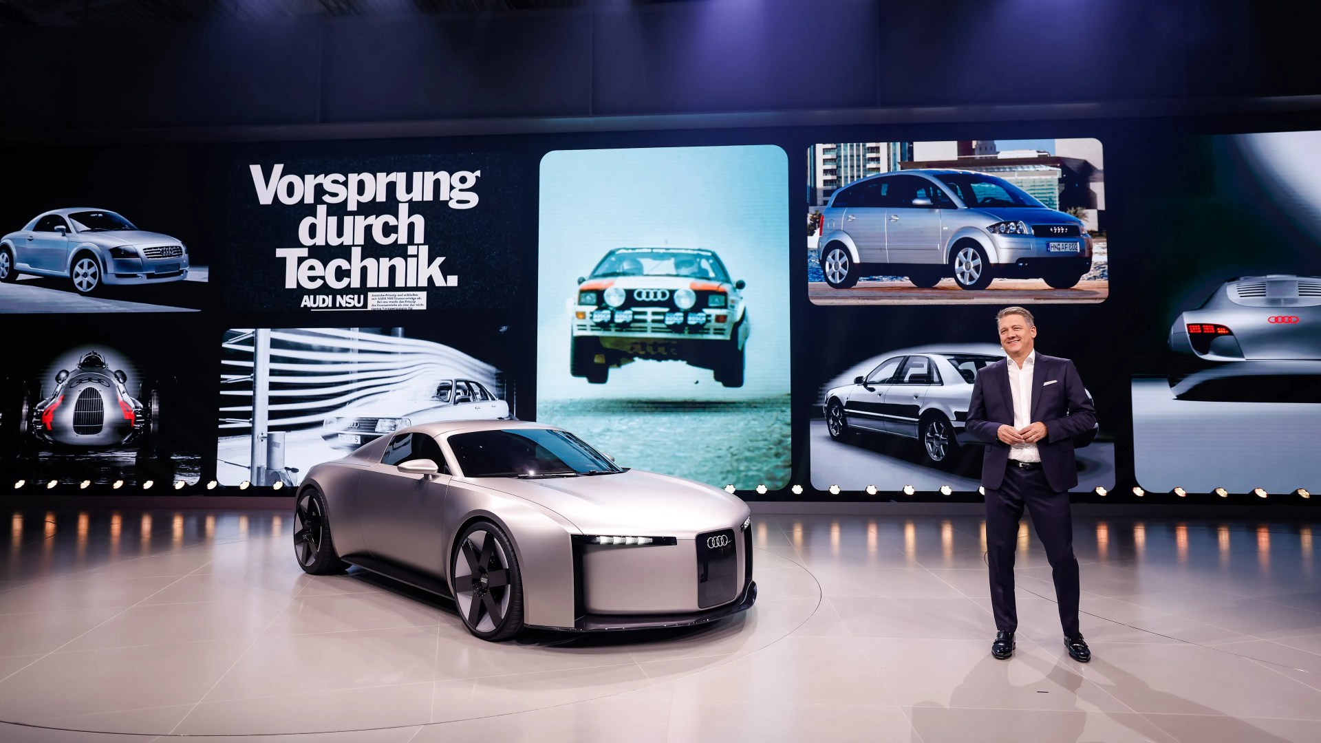 Silver futuristic Audi concept car on a showroom floor with a man in a suit and a large screen showing vintage Audi cars and the text "Vorsprung durch Technik.