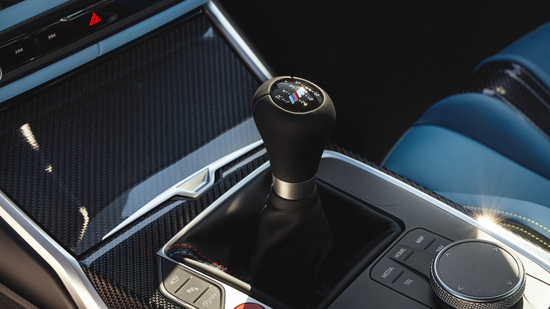 Black leather manual gear shift knob with BMW M logo in a car interior with carbon fiber trim and blue leather seats.