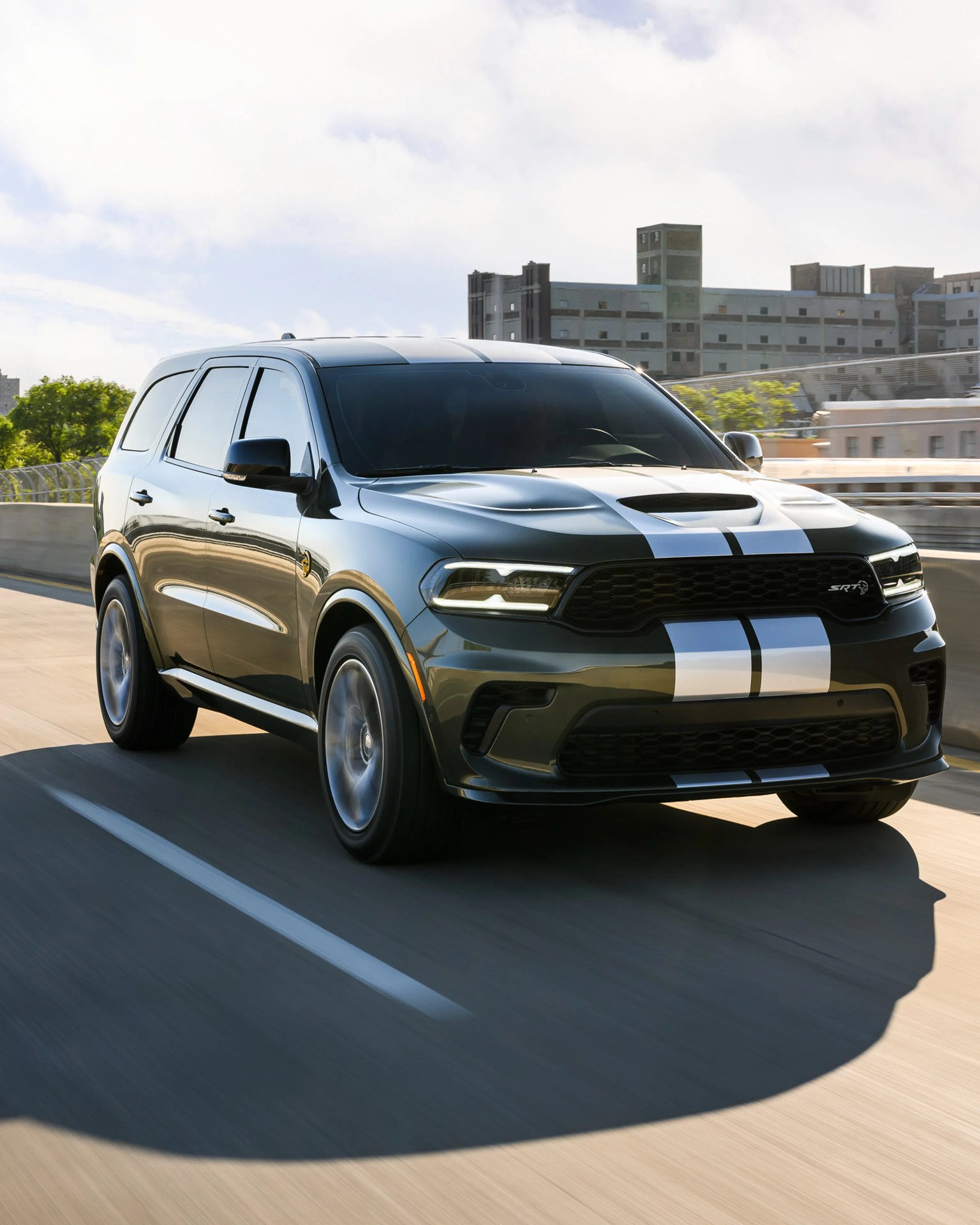 Dark green Dodge Durango SRT SUV with white racing stripes driving on a city highway.