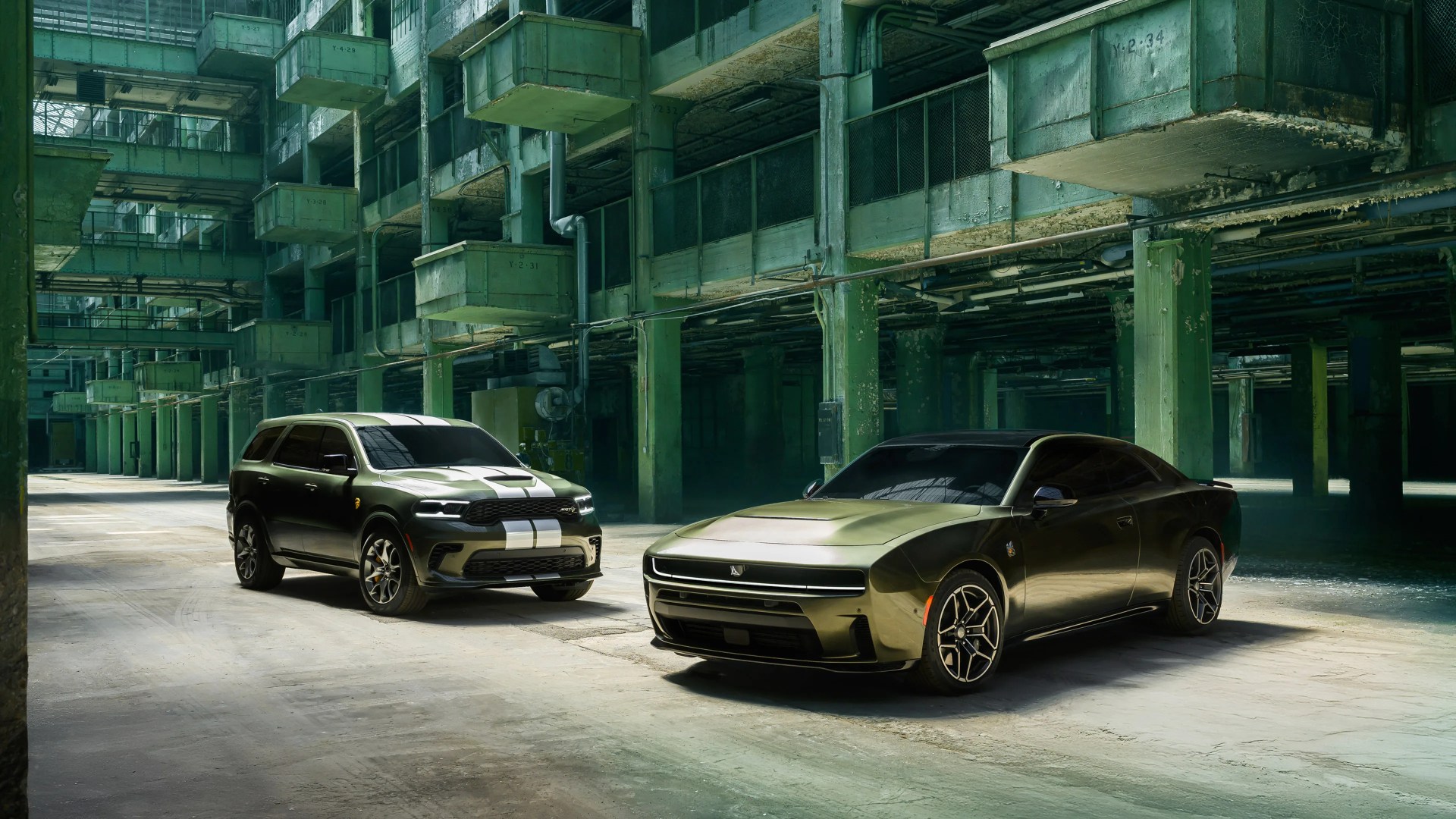 Two olive green Dodge vehicles, a muscle car and an SUV with white racing stripes, parked inside an industrial green-painted warehouse.