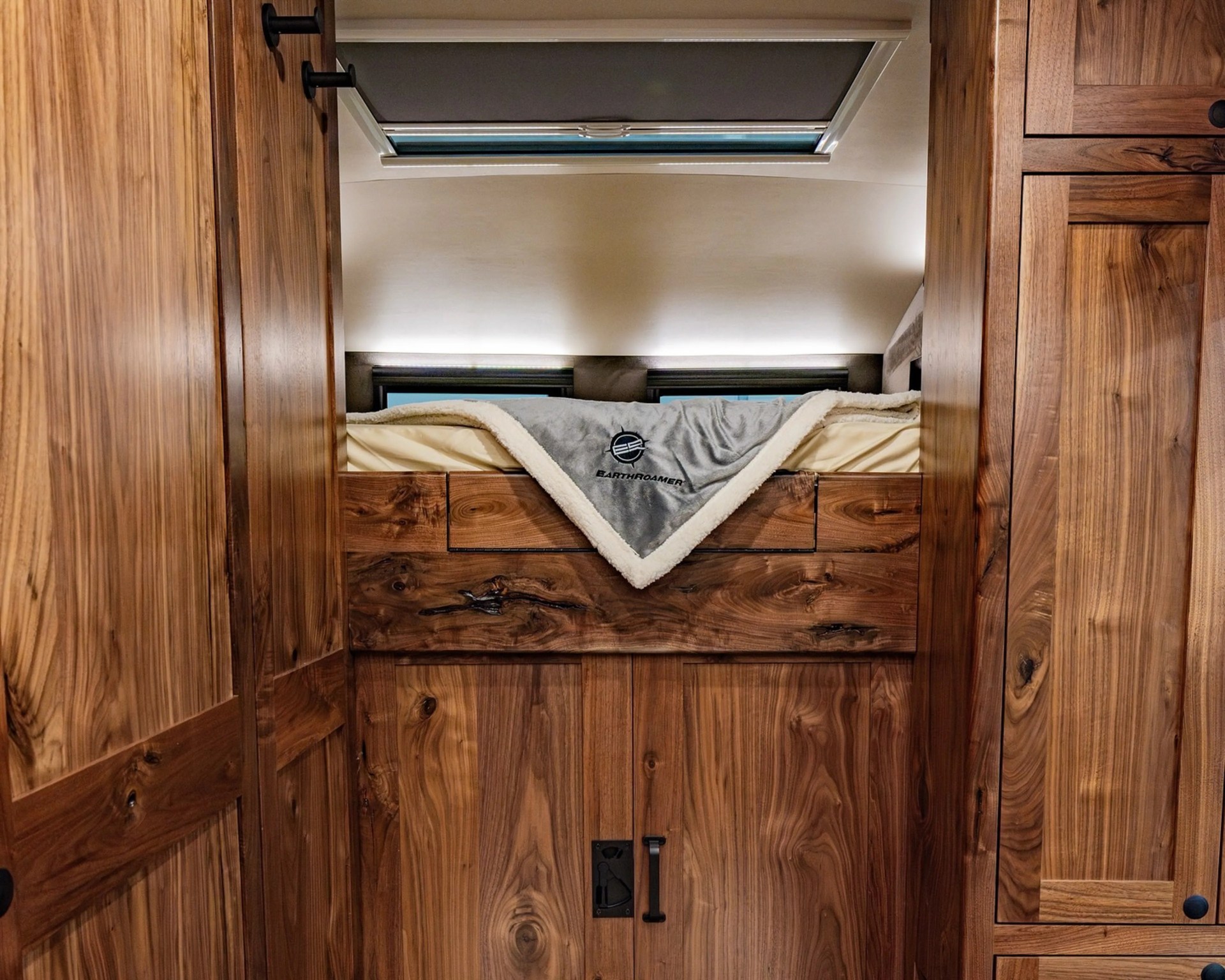 Wood-paneled RV interior with a raised bed featuring a gray EarthRoamer blanket and storage cabinets below.