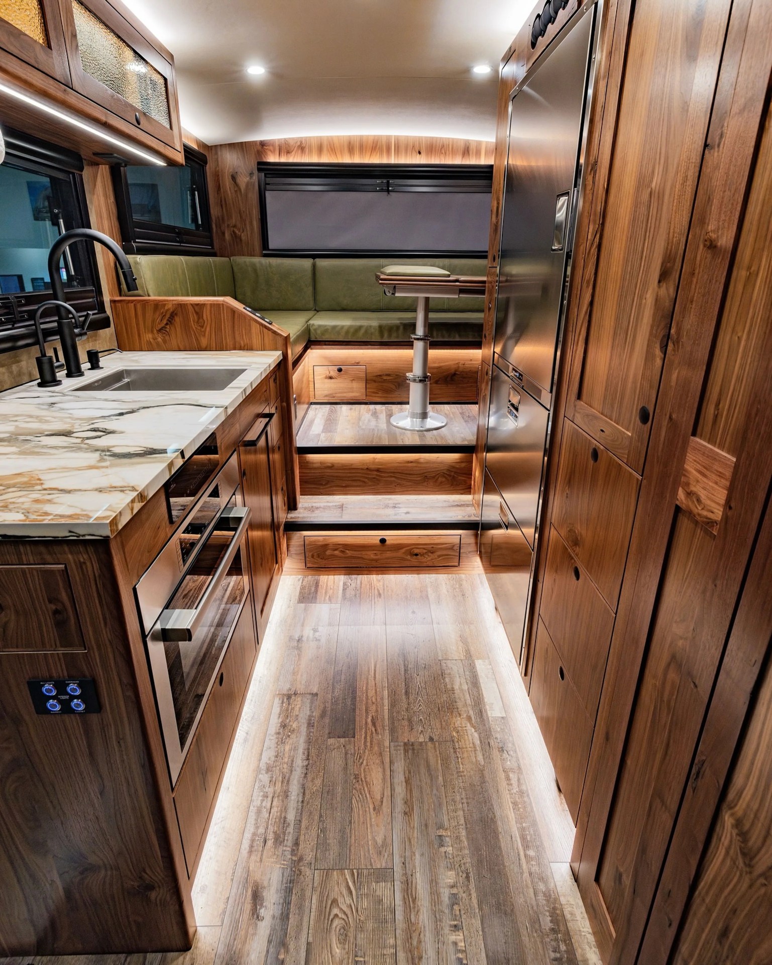 Compact RV kitchen and dining area with wood cabinetry, marble countertop, stainless steel appliances, and green cushioned seating.
