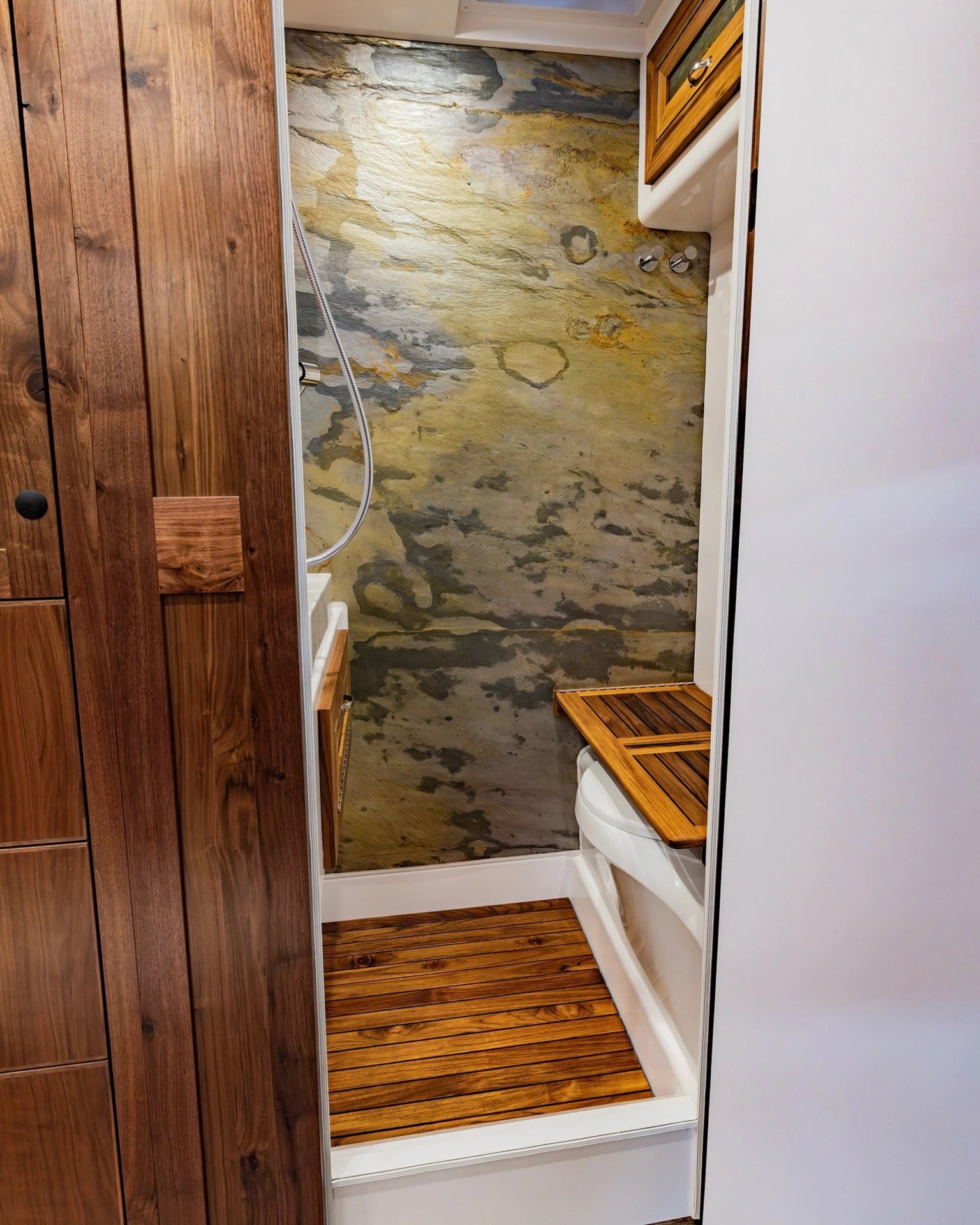 Compact shower area with wooden slat floor and bench, stone-textured wall, and wooden cabinetry.