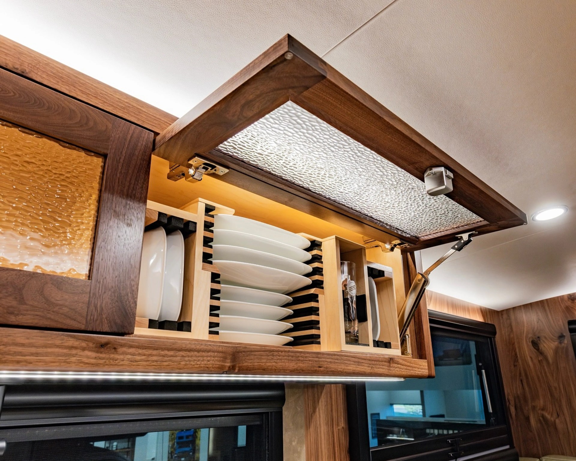 Wooden kitchen cabinet with textured glass door open, showing organized white plates and glass inside.