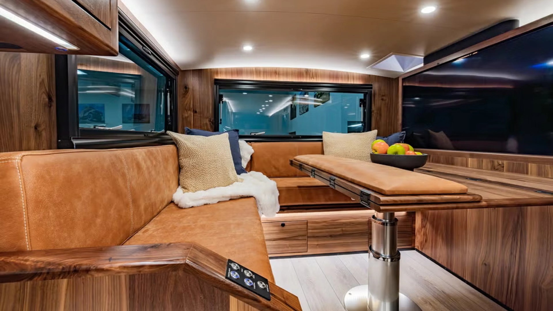 Interior of a camper van with tan leather L-shaped seating, wood paneling, a foldable table, and a bowl of fruit.