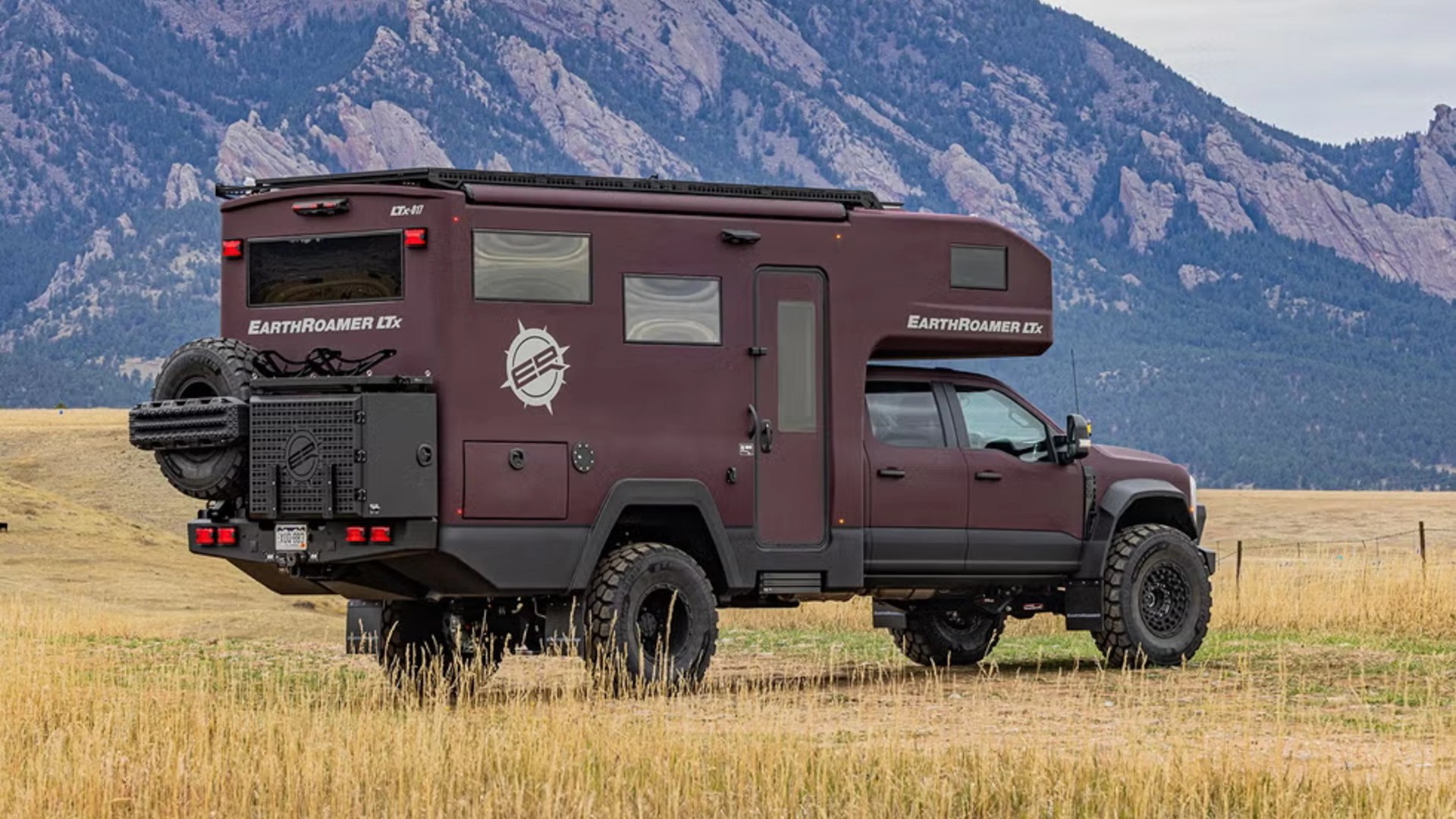Maroon EarthRoamer LTx off-road camper truck parked in a grassy field with mountains in the background.