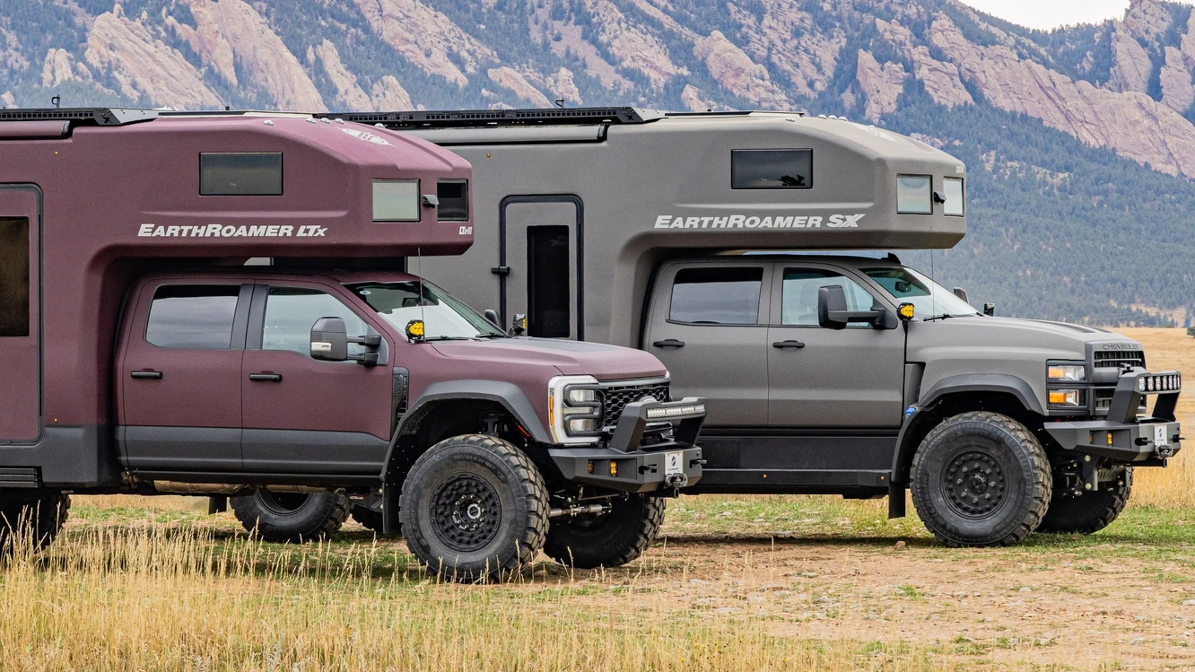 Two EarthRoamer expedition vehicles, one maroon EarthRoamer LTx and one gray EarthRoamer SX, parked on a grassy field with mountains in the background.