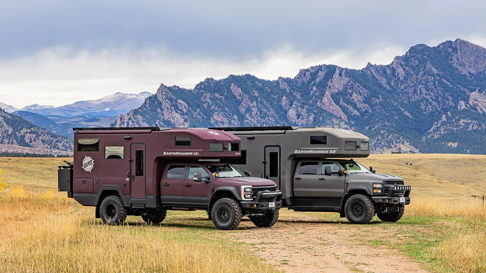 Two EarthRoamer off-road expedition trucks, one maroon and one gray, parked on a grassy plain with mountains in the background.