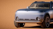 Front view of a matte silver electric SUV with LED headlights and rugged off-road tires on sand dunes.