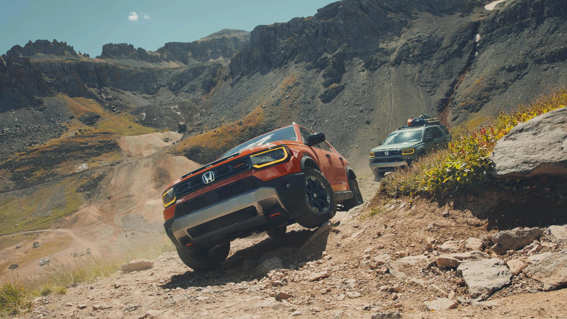 Orange Honda SUV with yellow headlights off-roading on rocky mountain trail with another green SUV behind.
