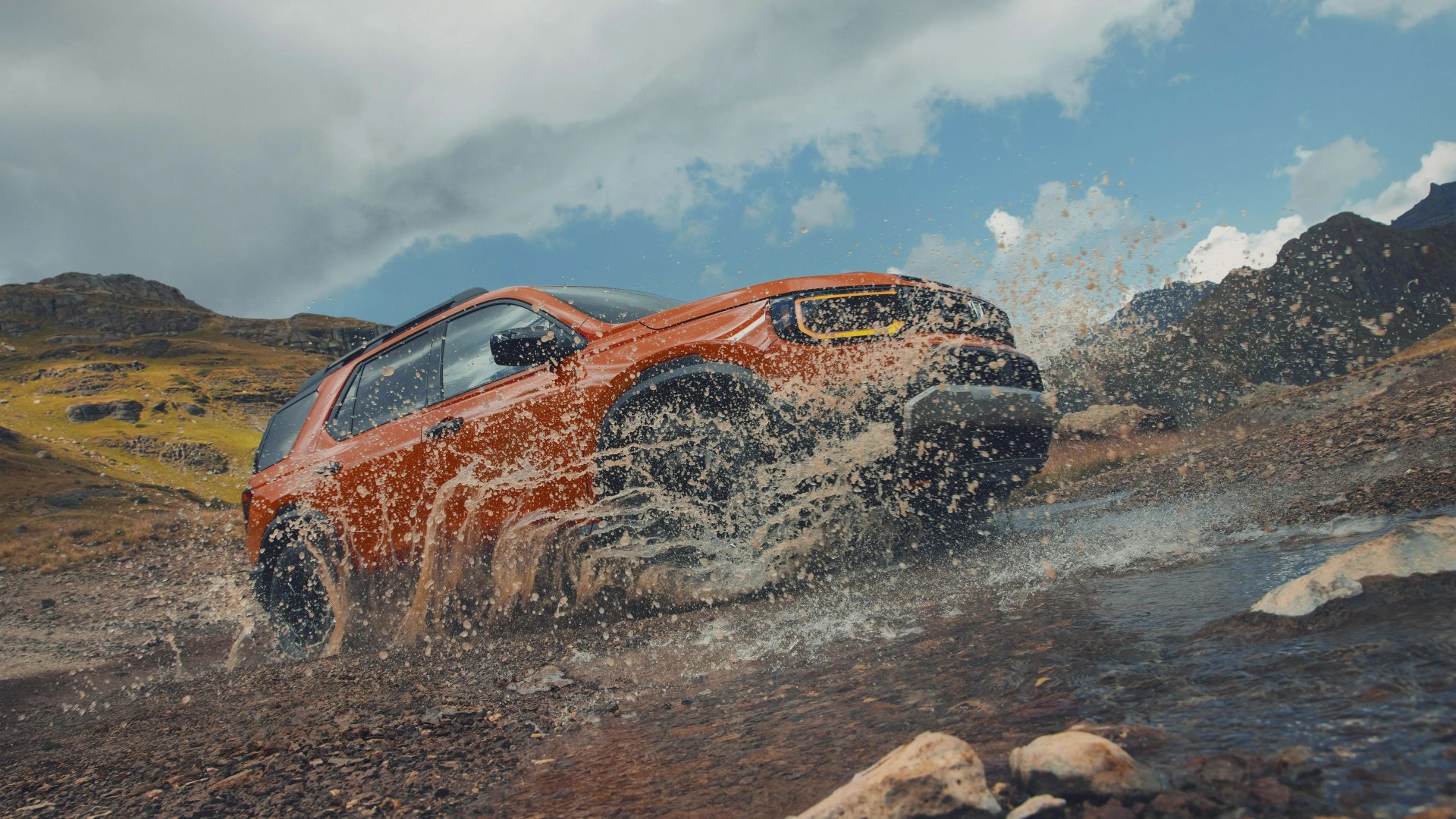 Orange off-road vehicle splashing through a shallow rocky stream in a mountainous landscape.