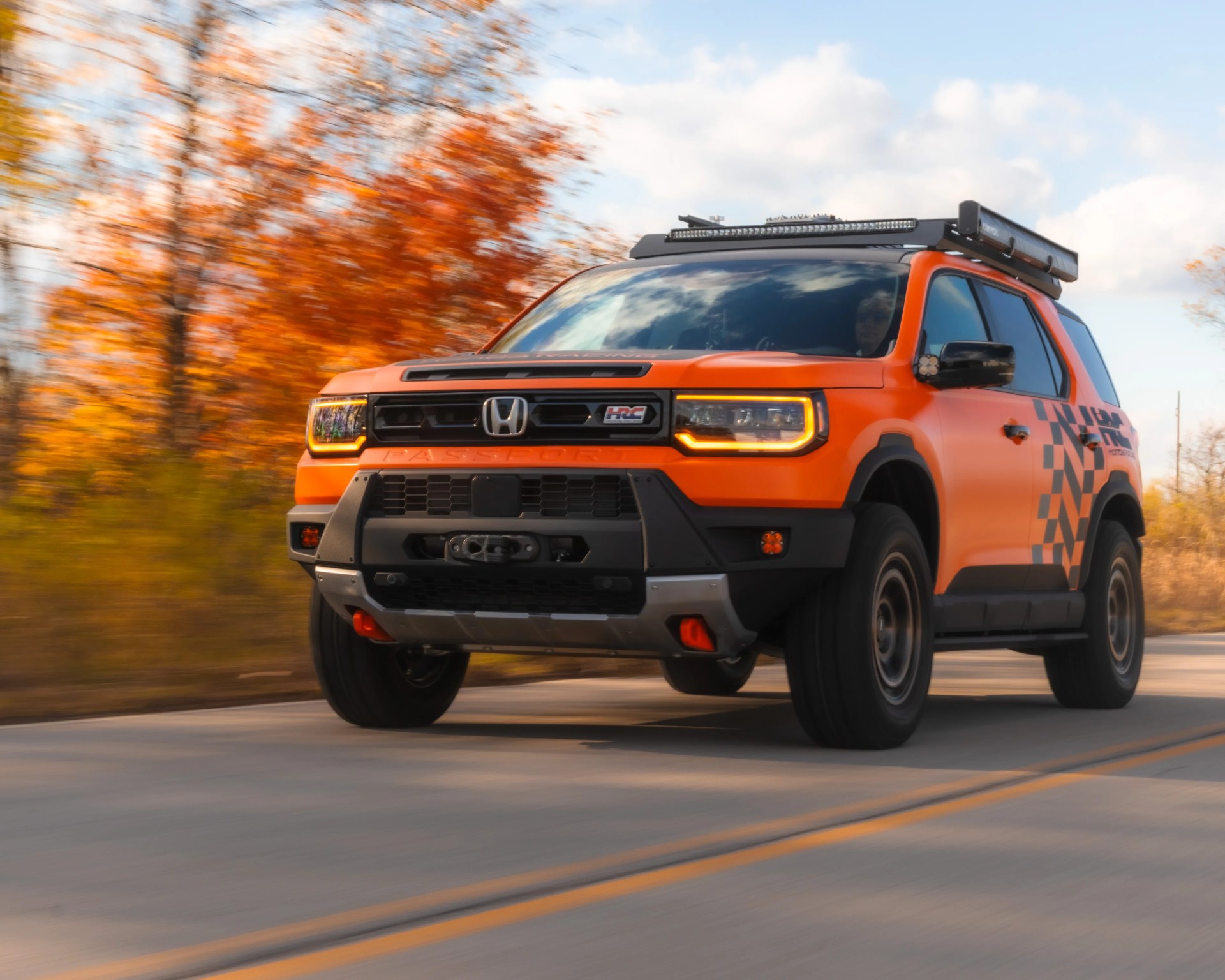 Orange Honda SUV with black roof rack and checkered side decals driving on a road with autumn trees in the background.