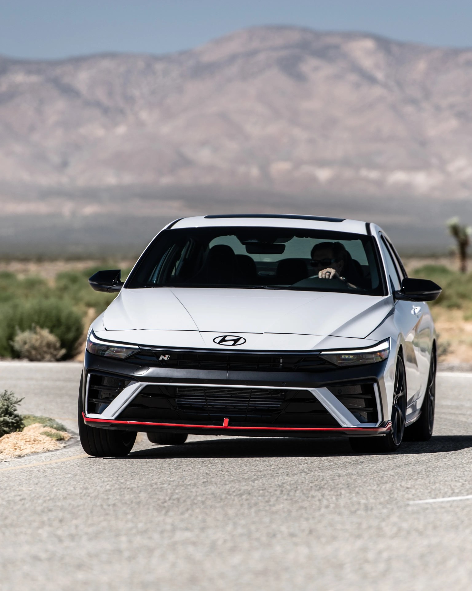 White Hyundai sedan with black and red front grille details driving on a desert road with mountains in the background.