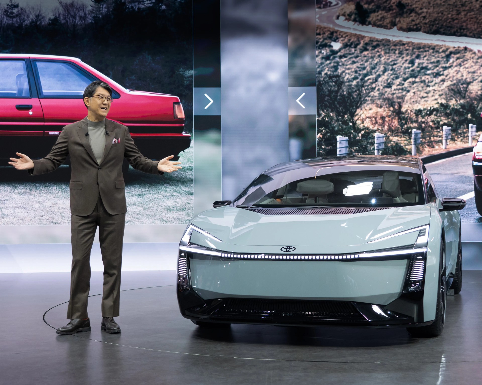 Man in brown suit presenting a light green futuristic Toyota car with LED headlights on a showroom floor.