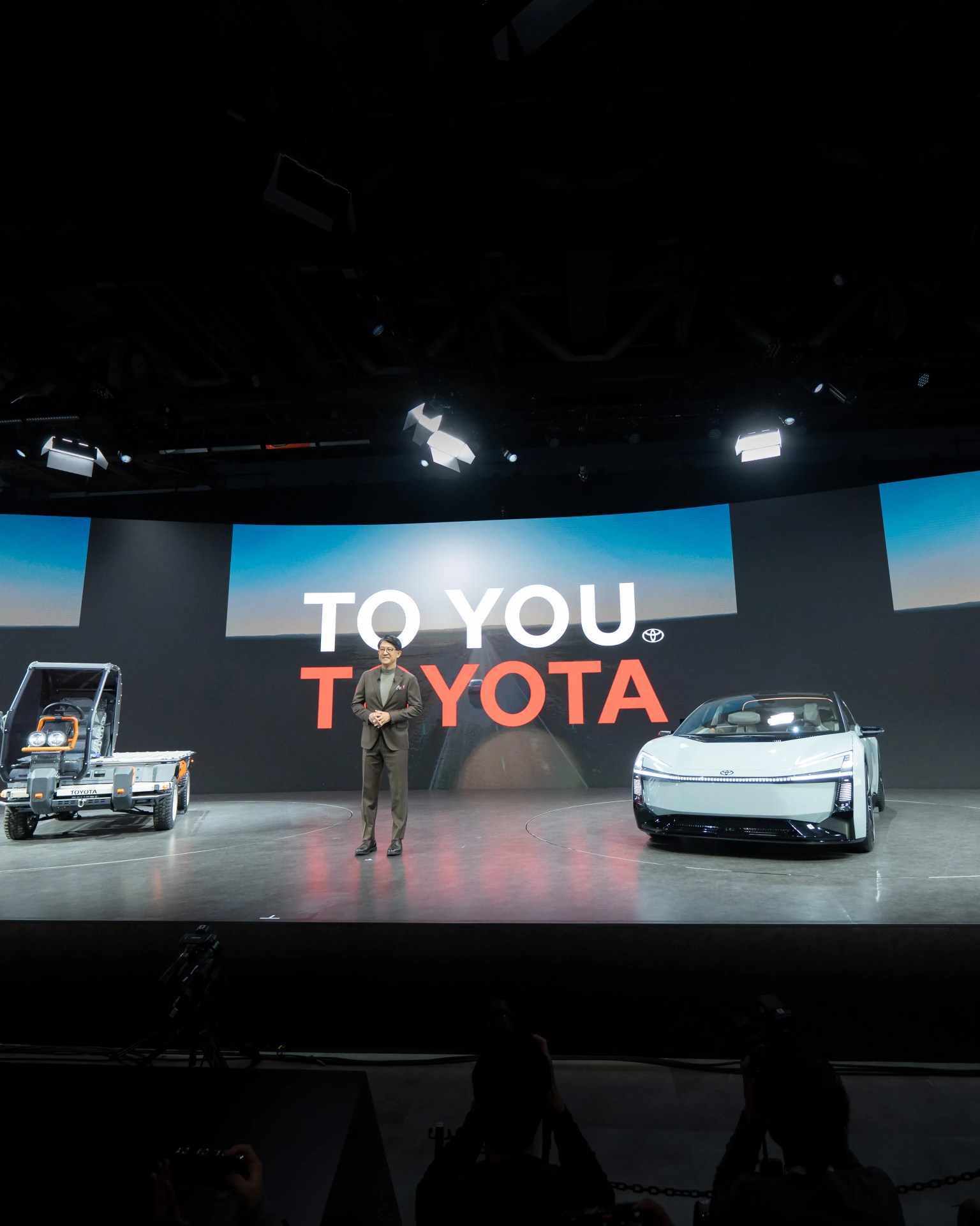 Man in a suit standing on stage between a futuristic white Toyota car and a small utility vehicle with "TO YOU TOYOTA" displayed on a large screen behind.