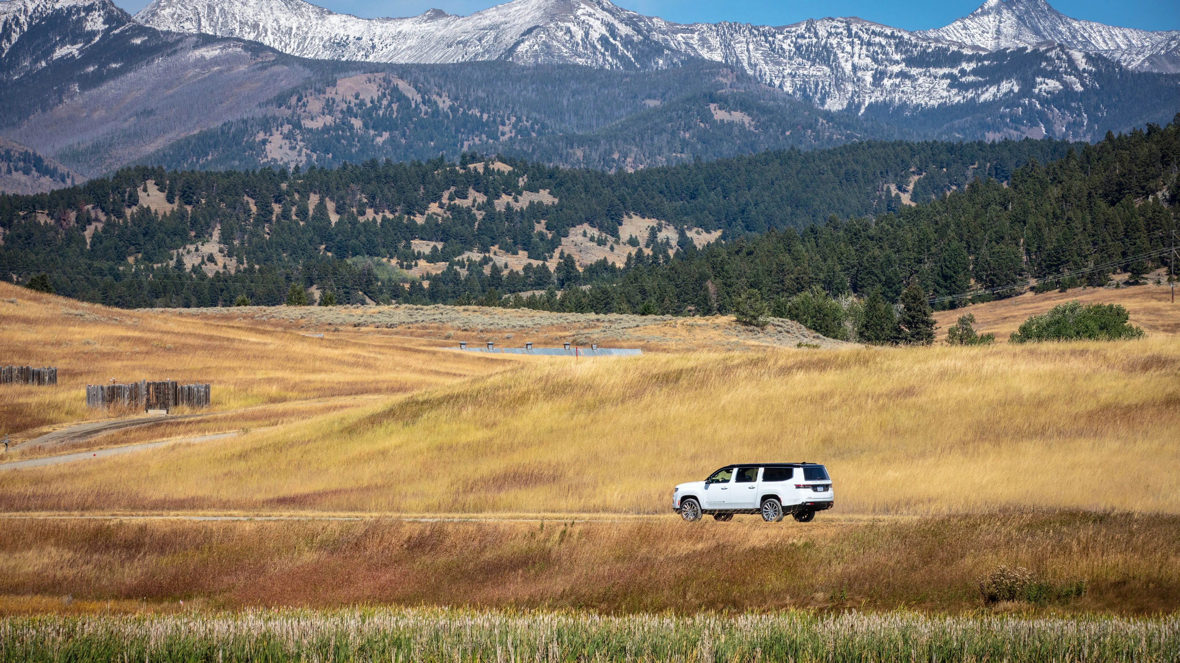 White SUV driving on a dirt road through golden grass fields with forested hills and snow-capped mountains in the background.