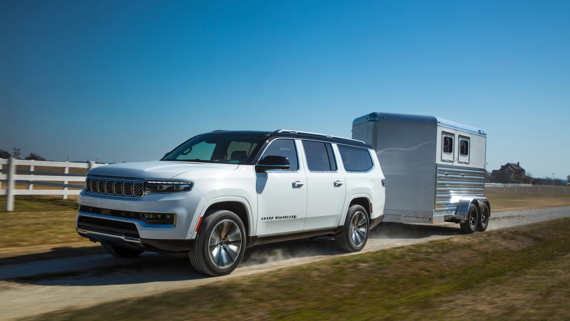 White SUV with black roof towing a silver two-axle trailer on a dirt road with a white fence in the background.