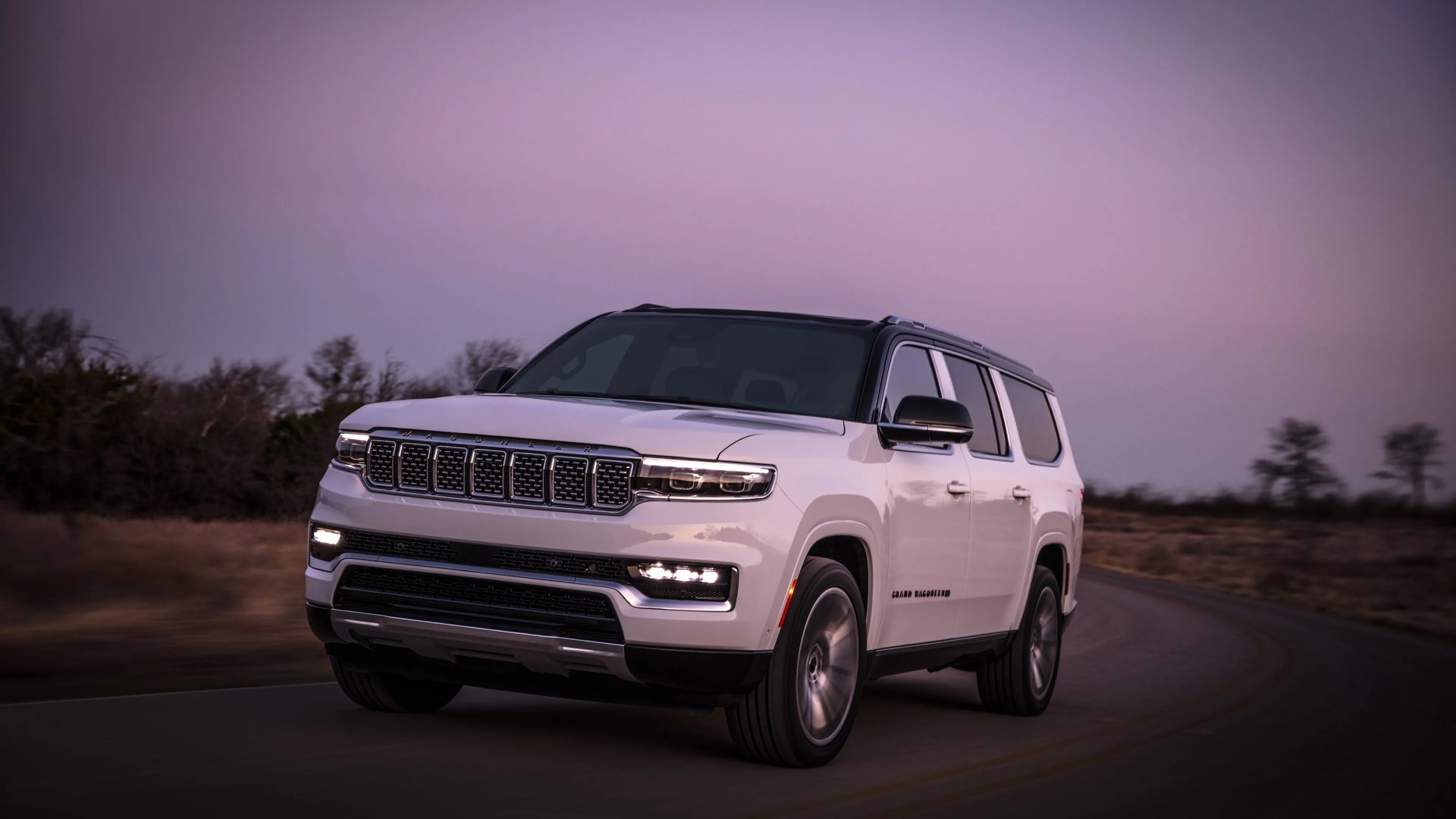 White Jeep Grand Wagoneer SUV with black roof driving on a curved road at dusk.