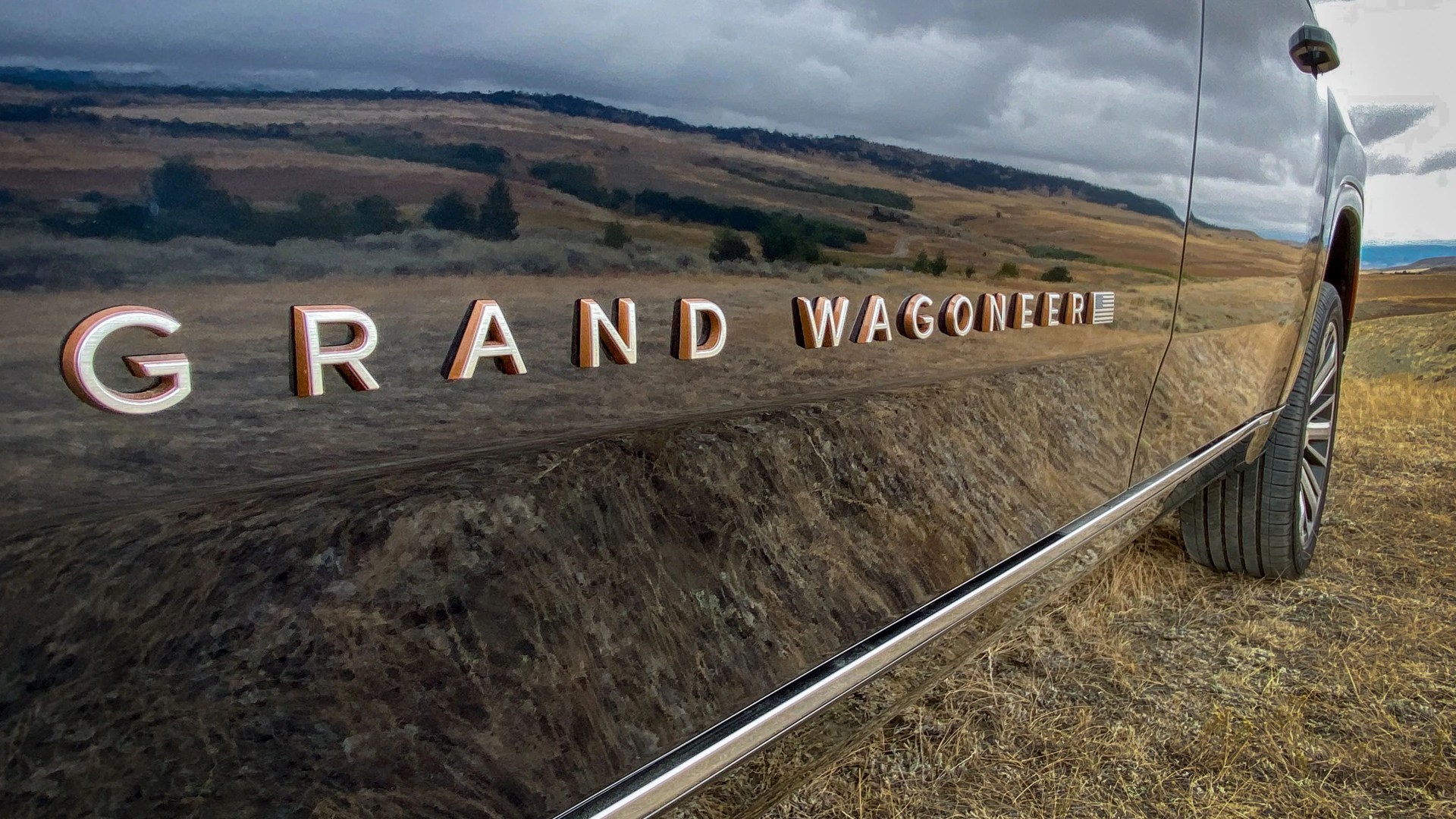 Glossy black vehicle side panel with "GRAND WAGONEER" emblem and a rear tire on dry grass.