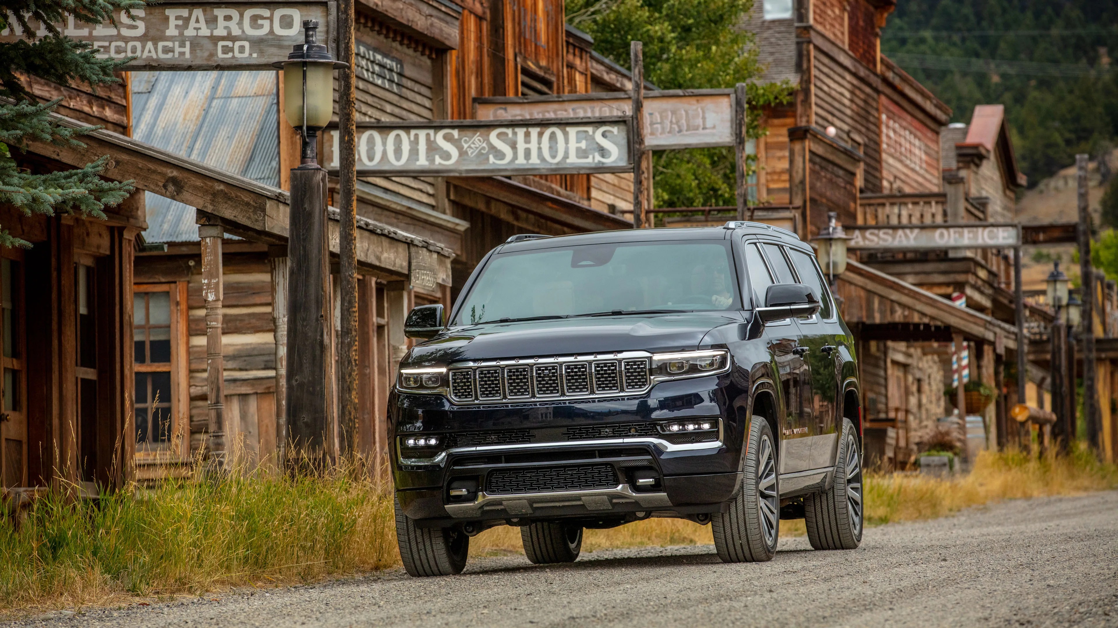 Black Jeep Wagoneer SUV parked on a gravel road in front of rustic wooden buildings with vintage signs.