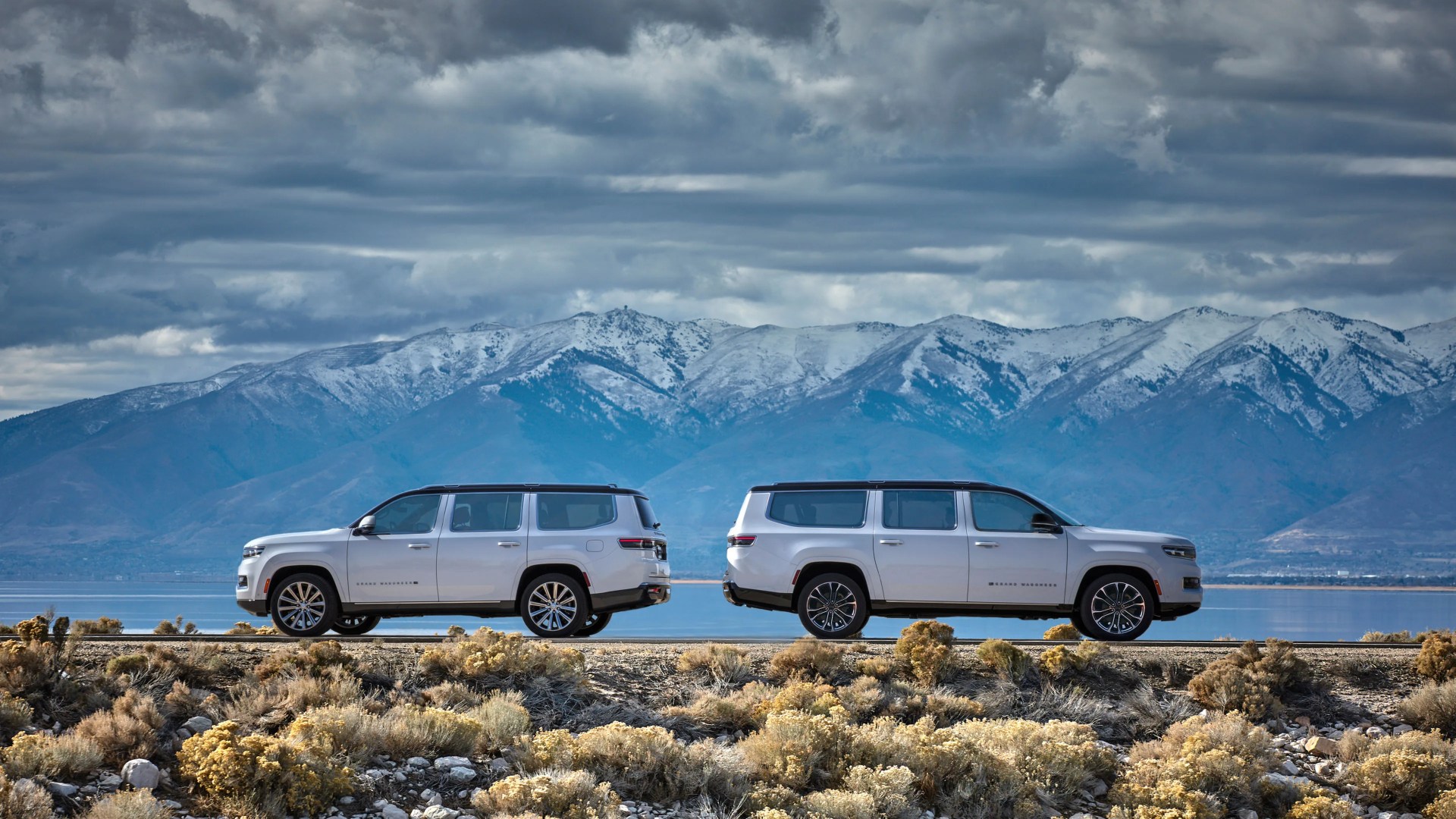Two silver SUVs parked side by side on a road with snow-capped mountains and a cloudy sky in the background.