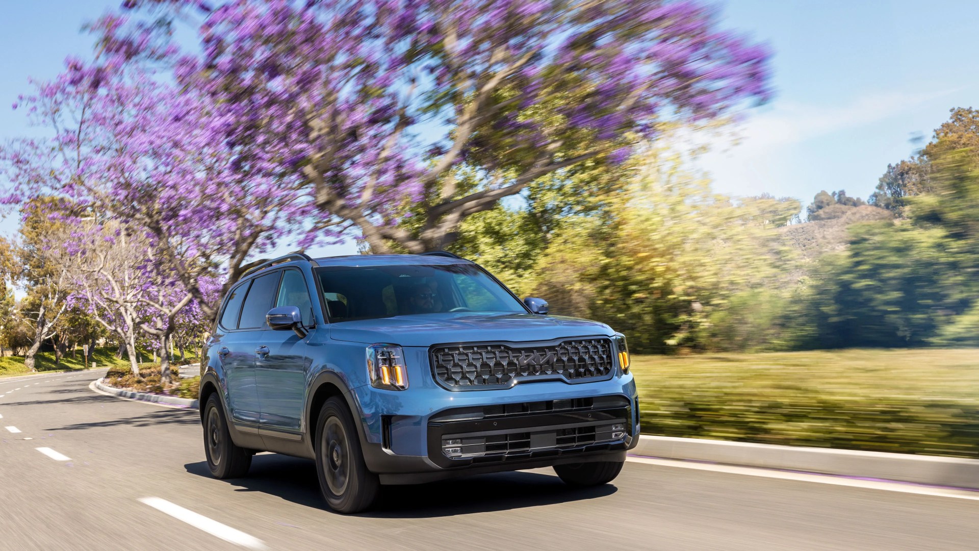 Blue Kia SUV driving on a road with purple-flowered trees in the background.
