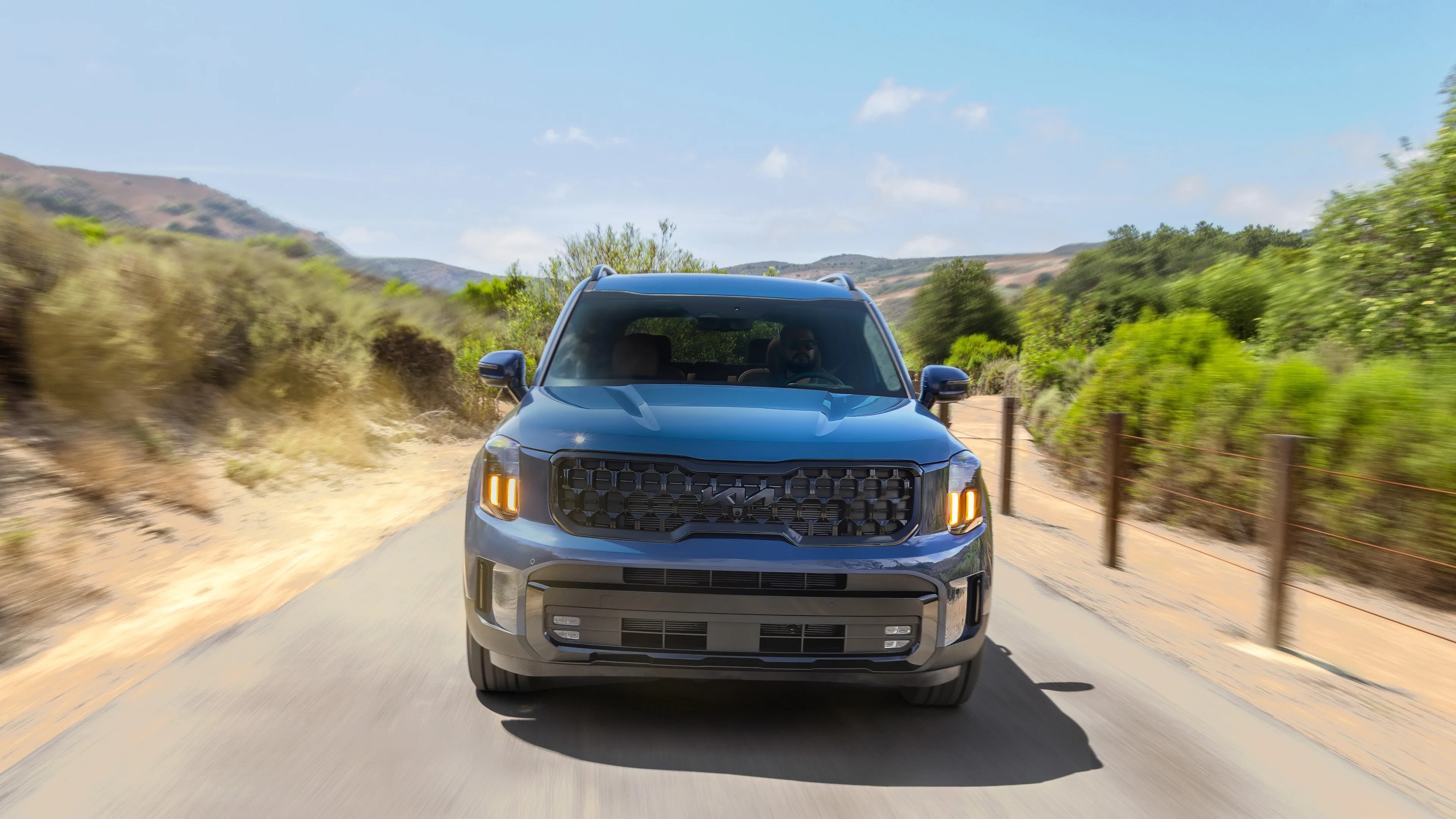 Blue Kia SUV with illuminated amber headlights driving on a rural road with greenery and hills in the background.