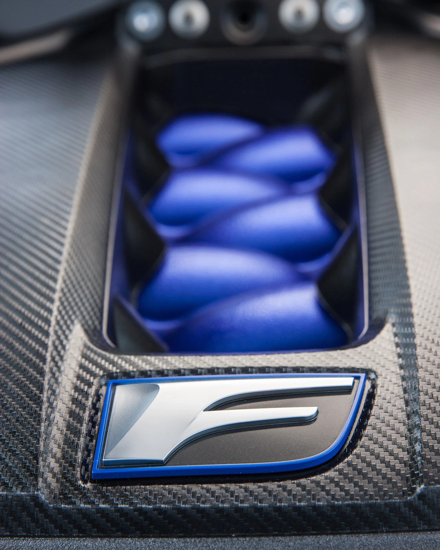 Close-up of a carbon fiber car part with a silver and blue "F" logo and blue leather detail in the background.