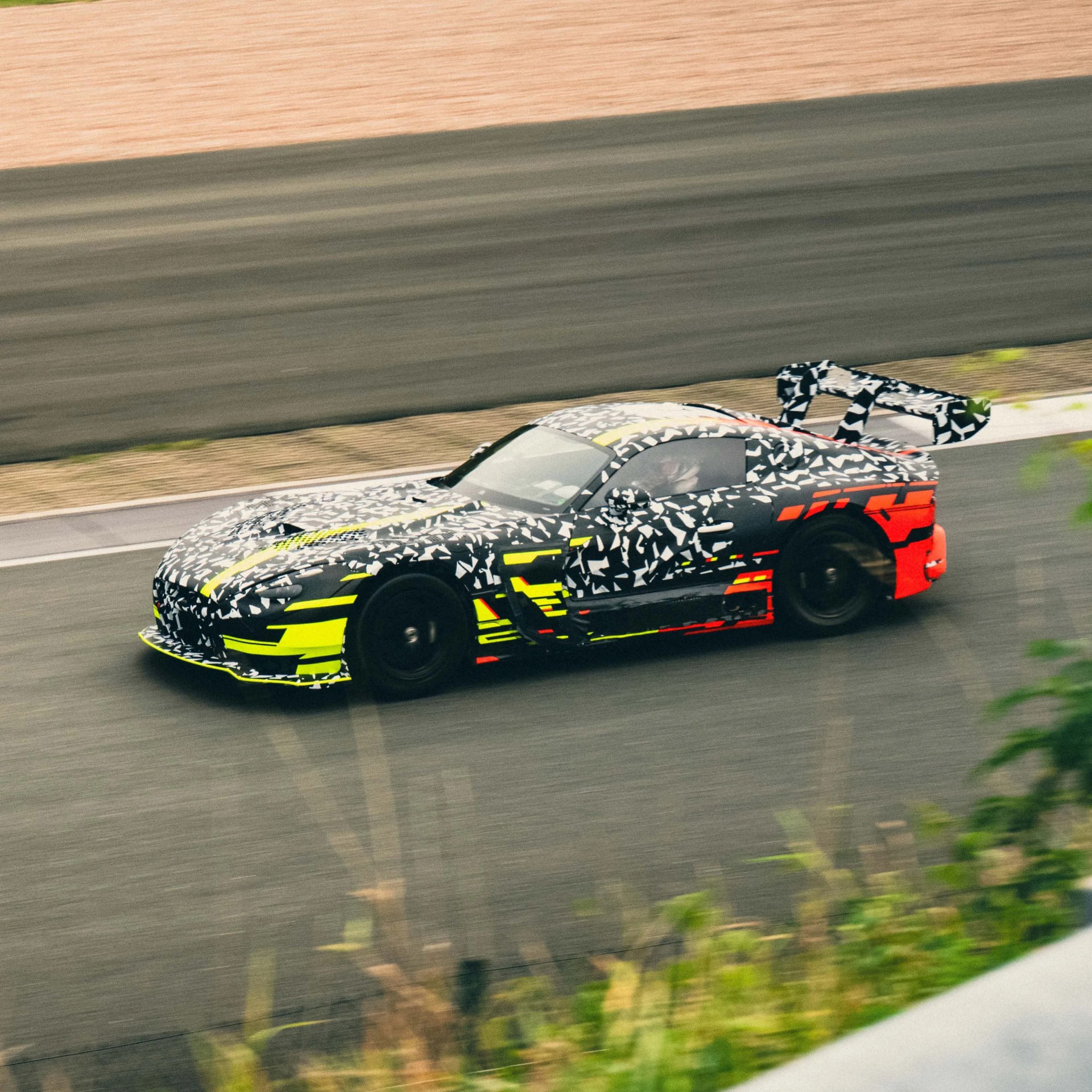 Black race car with white, yellow, and red camouflage pattern and large rear wing on a racetrack.