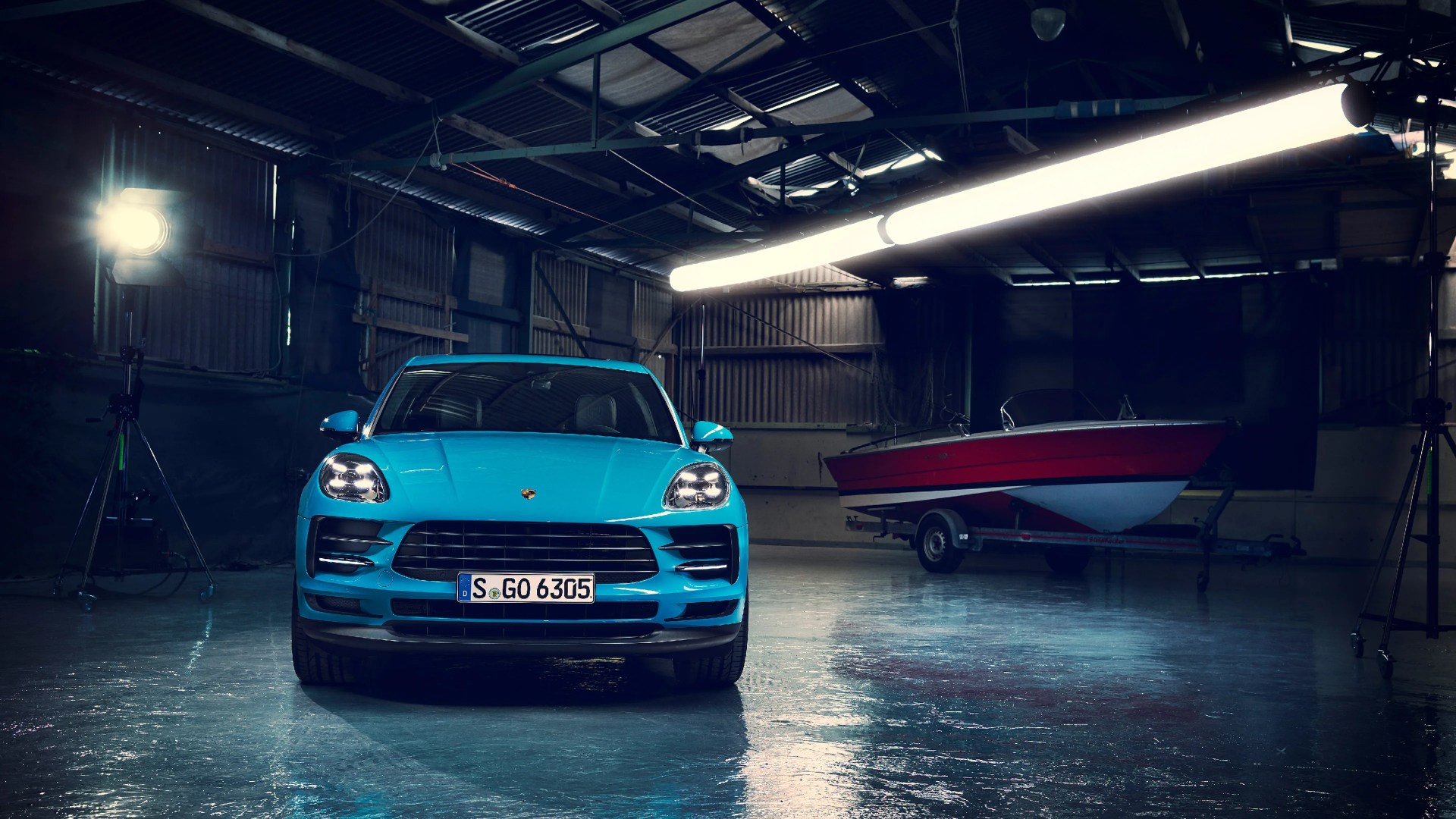 Bright blue Porsche SUV with headlights on in a dimly lit industrial garage with a red and white boat on a trailer in the background.