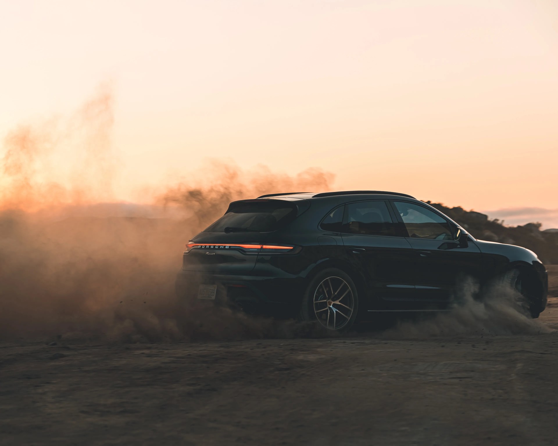 Black Porsche Macan SUV kicking up dust on a dirt road at sunset.