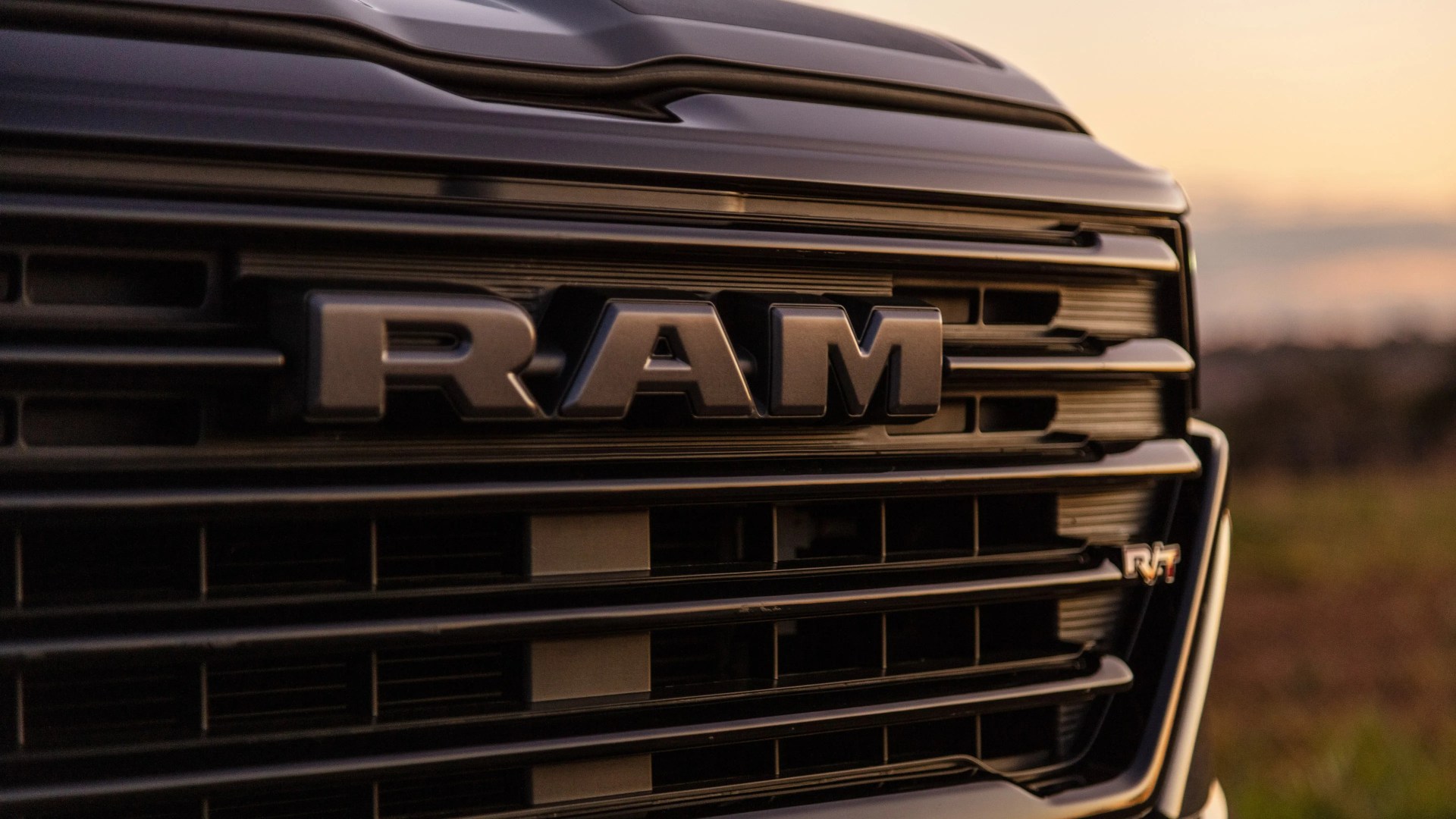 Close-up of a black RAM truck grille with bold RAM lettering and an R/T badge at sunset.