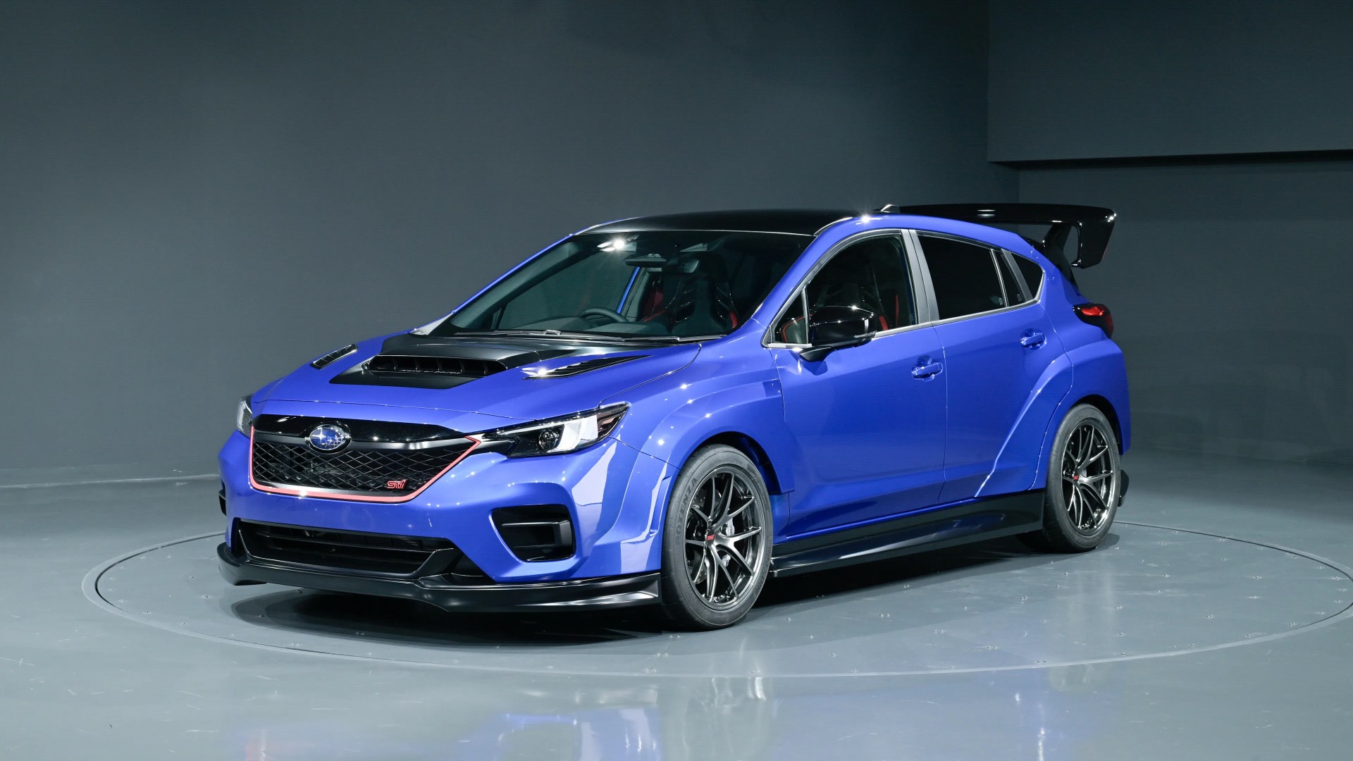 Blue Subaru hatchback with black roof, hood scoop, wide fenders, and large rear spoiler in a studio.